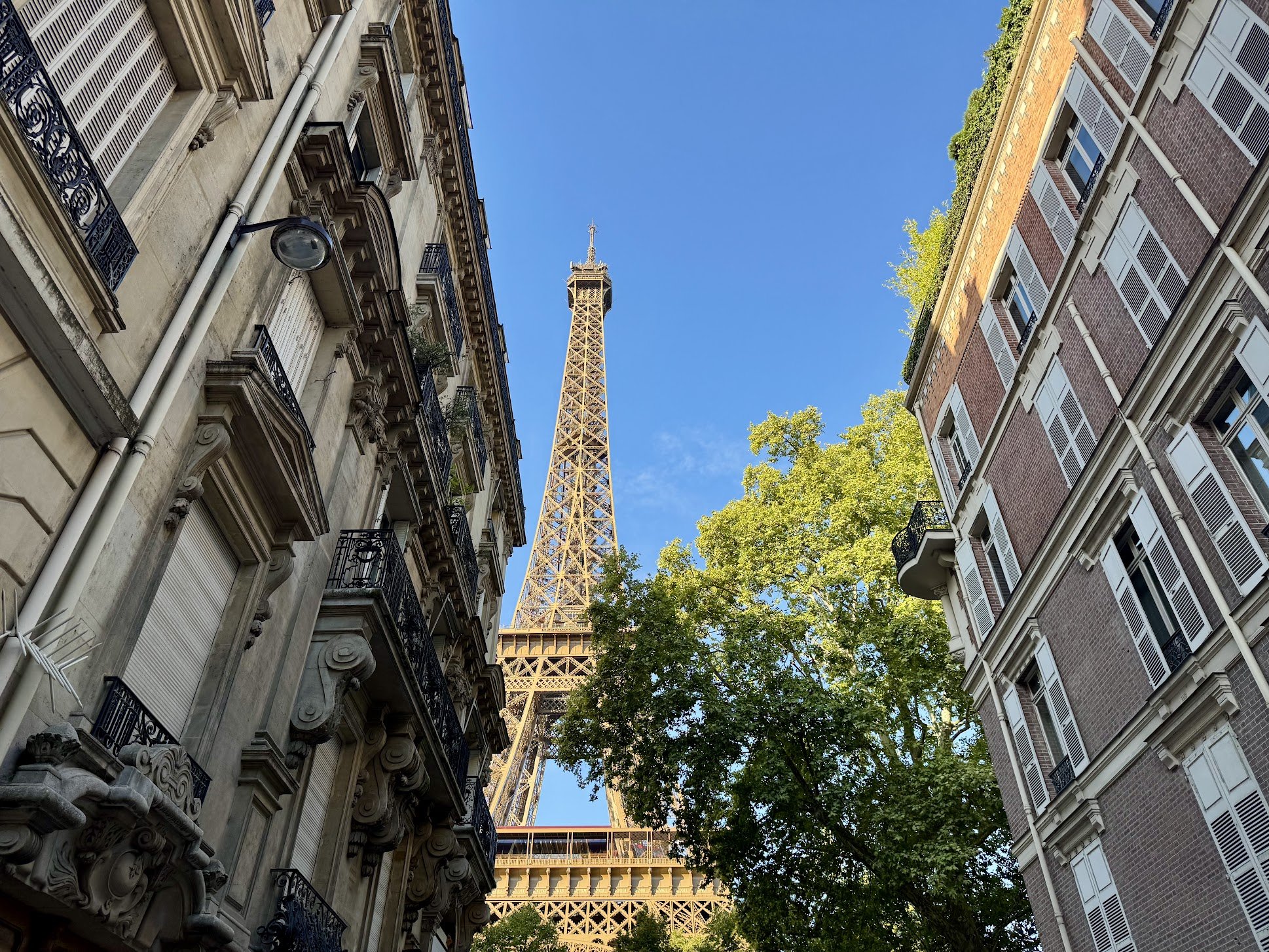 View of the Eiffel Tower in Paris, France, seen from a narrow street framed by classic European architecture, with balconies, white shutters, and leafy green trees against a clear blue sky.