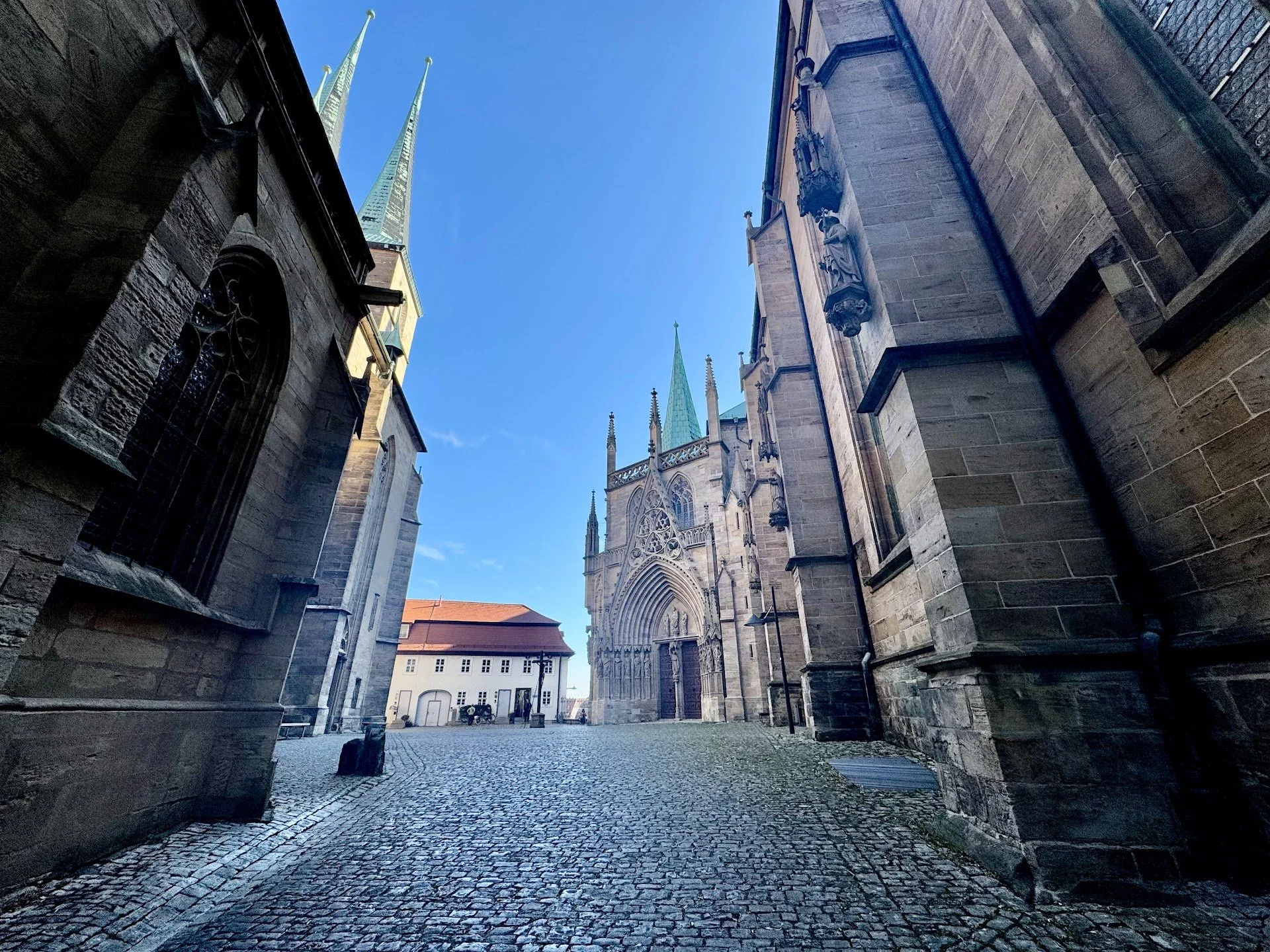A view of a historic church with gothic architecture, large arched doorway, tall spires, and ornate sculptures on the stone facade, seen from a cobblestone courtyard under a clear blue sky.
