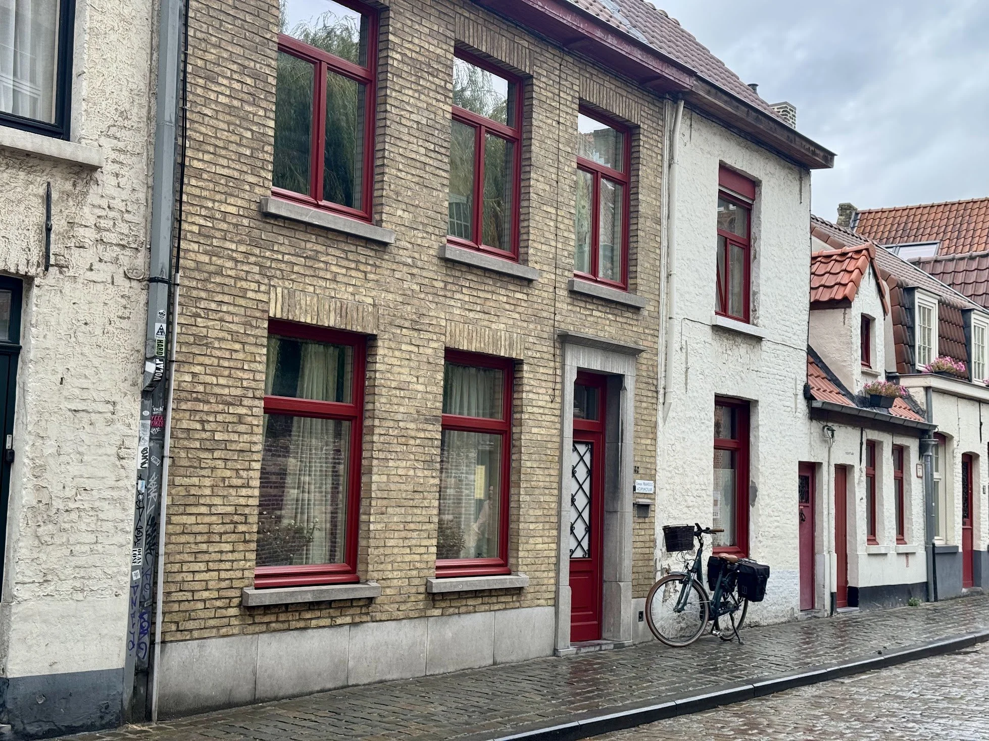Row of European-style buildings with brick and stucco facades, red window frames, and red-tiled roofs, street with cobblestones, and a bicycle parked outside.