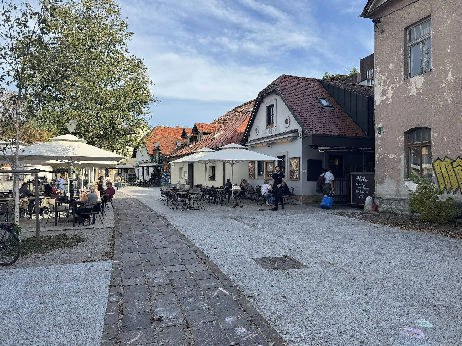 Outdoor cafe with tables and chairs, umbrellas, people sitting and walking, buildings with signs, trees, and a blue sky with clouds.