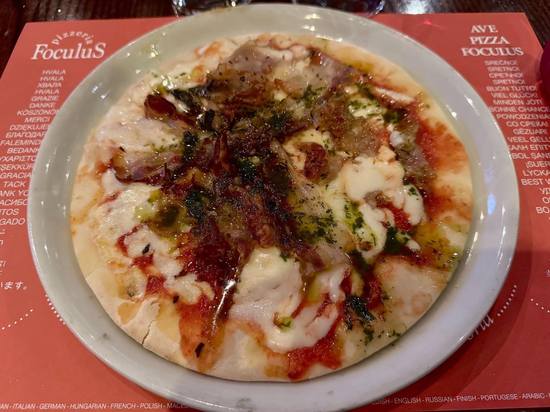 A pizza with cheese, tomato sauce, and various toppings including herbs and possibly bacon, served on a white dish on a red placemat.