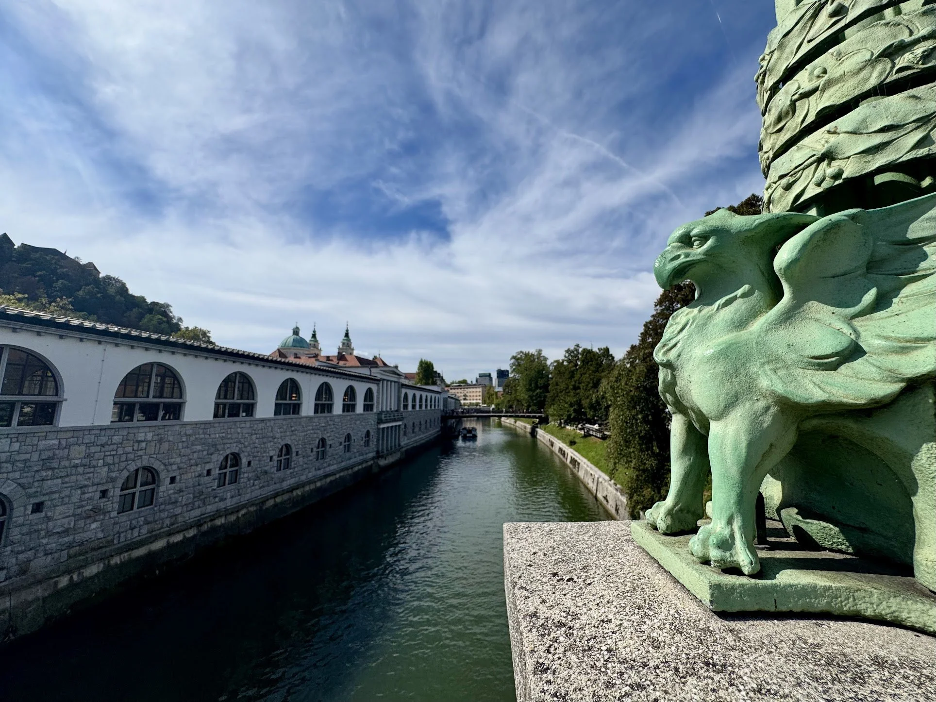 close-up of a green dragon sculpture on a bridge over a river, with a cityscape and two church steeples in the background under a partly cloudy sky.