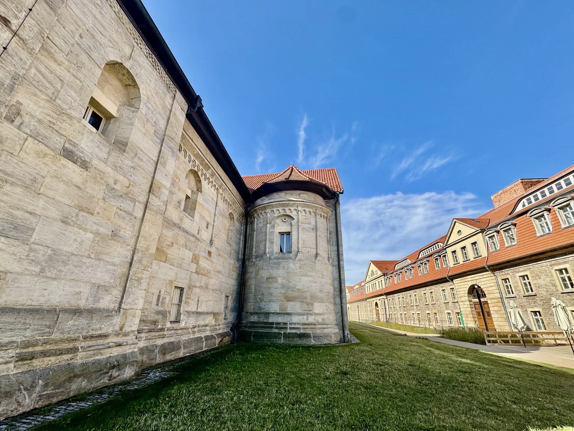A historic stone building with small arched windows and a turret, next to a row of modern buildings with red roofs under a blue sky.