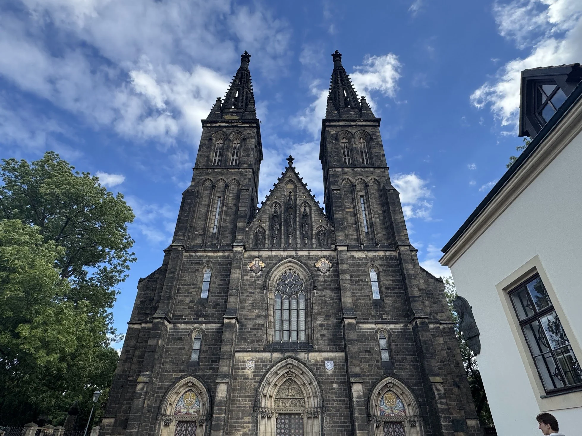 View of a large gothic-style church with two tall spires against a partly cloudy sky. The church is made of dark stone with intricate architectural details, stained glass windows, and some decorative elements. On the left, a tree with green leaves is visible. On the right, part of a white building and a person are seen.