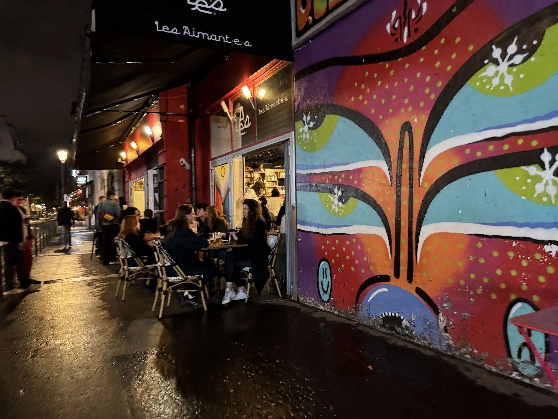 People sitting at outdoor tables outside a restaurant with a colorful mural wall at night.