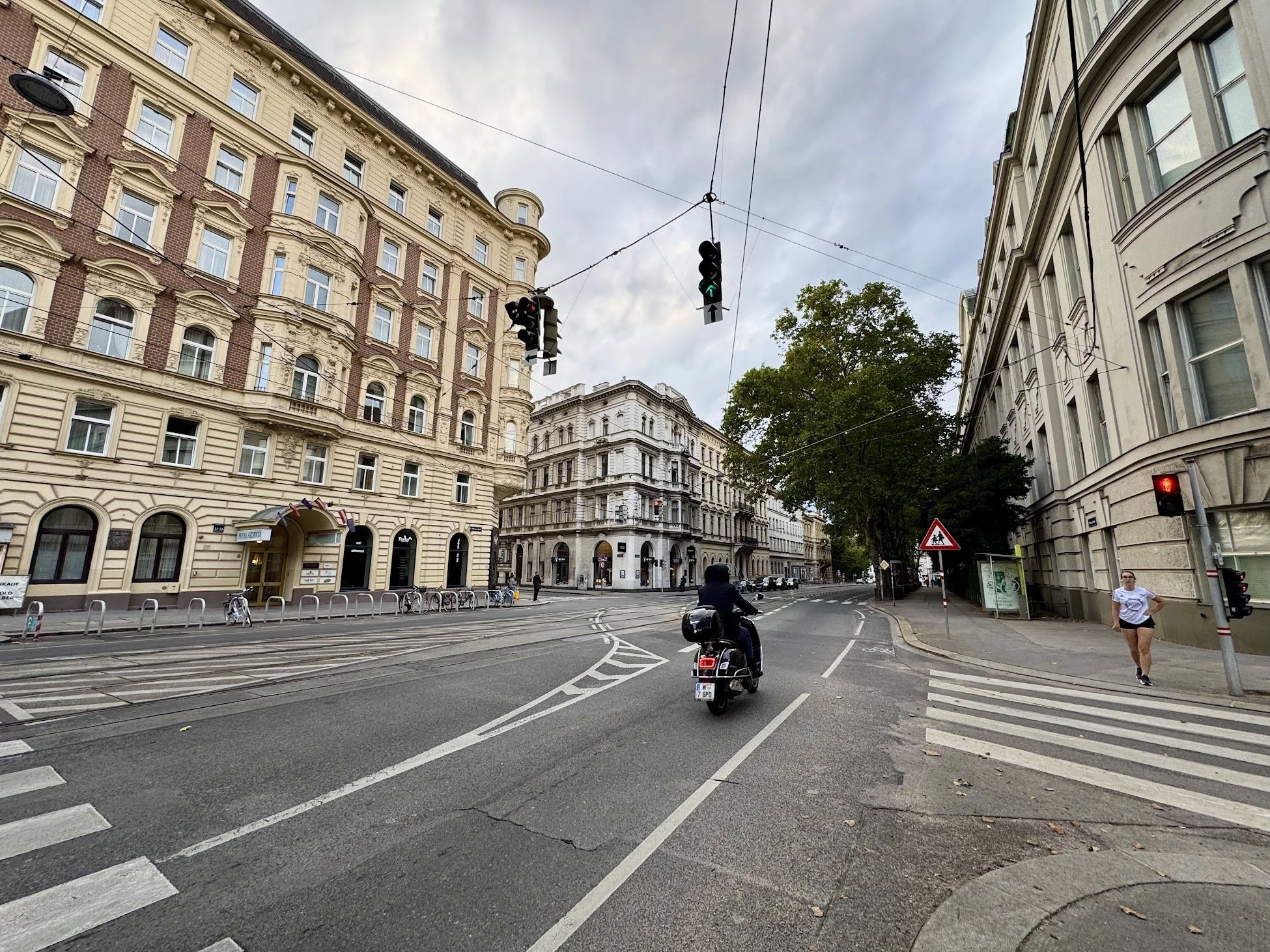 City street scene with tall historic buildings, a traffic light showing green, tram tracks, a person on a scooter, pedestrians crossing, and a large leafy tree in the background with a cloudy sky.