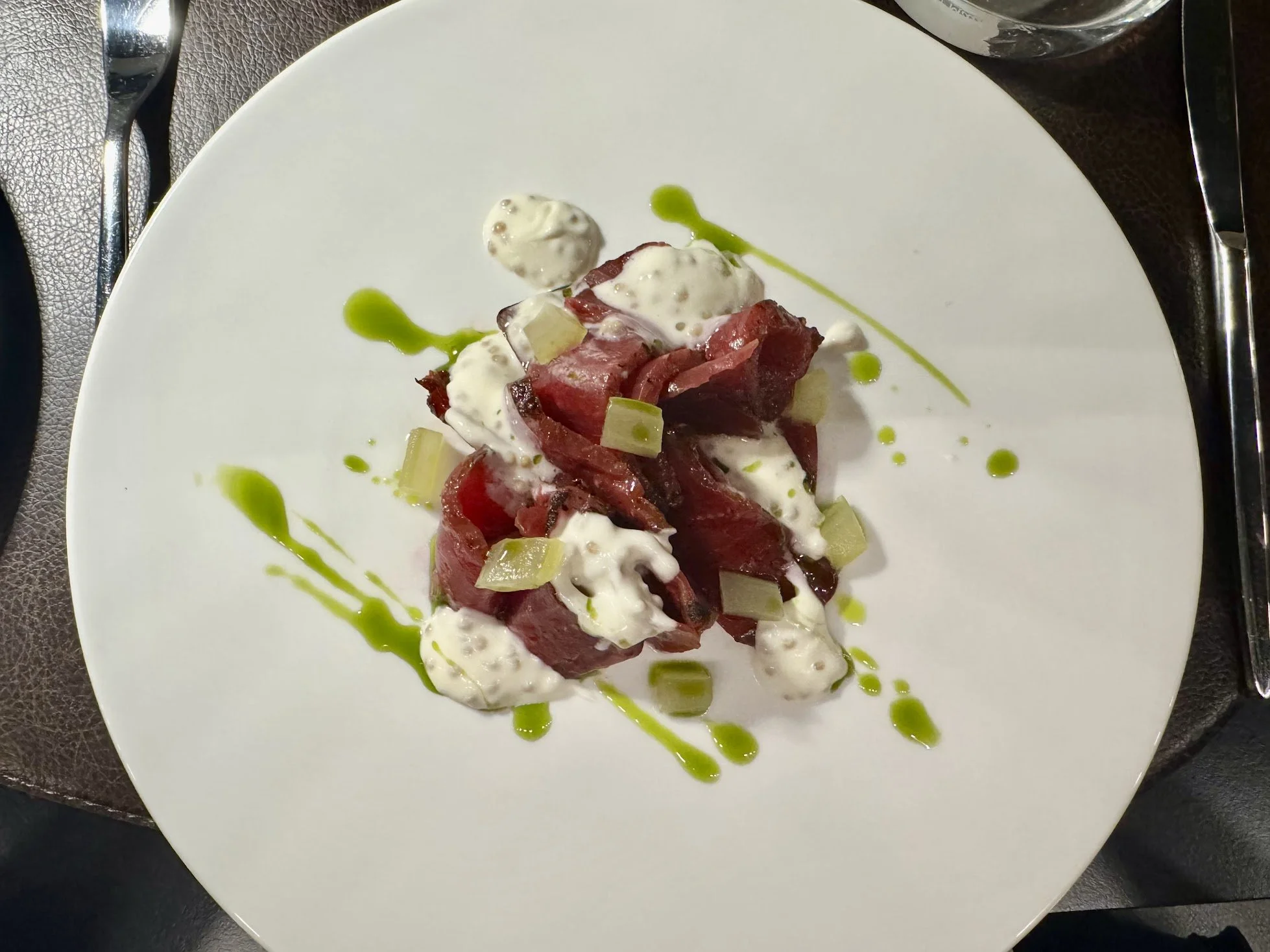 A plated dish featuring slices of cured meat topped with a creamy white sauce, garnished with chopped green vegetables and a drizzle of green oil, served on a white plate.