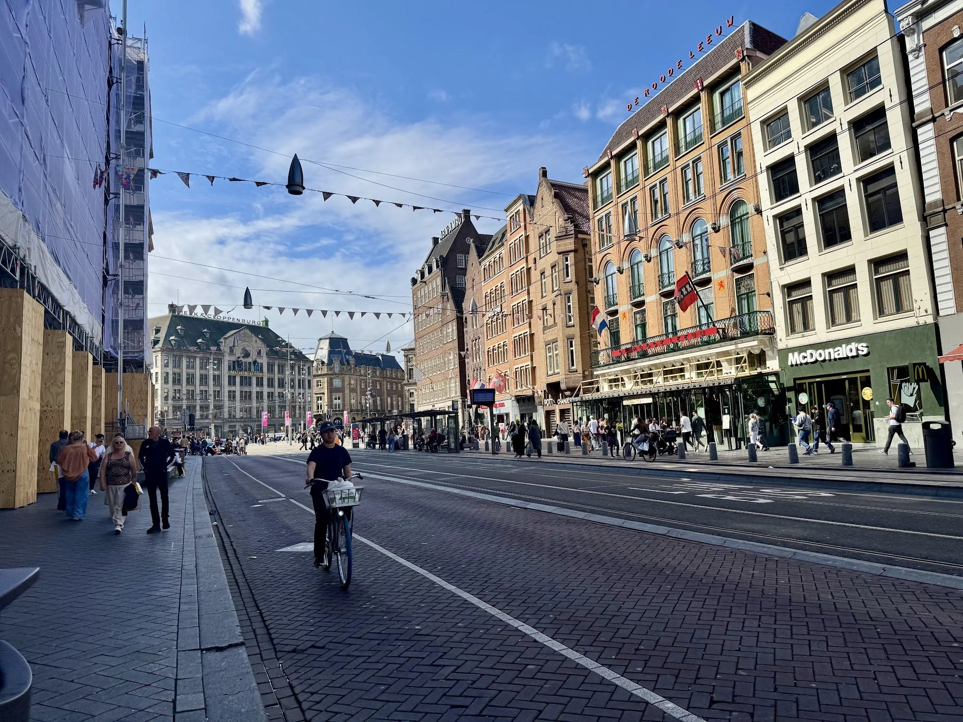 A bustling city street with people walking and biking, tram tracks on the road, and buildings with shops and apartments under a blue sky.