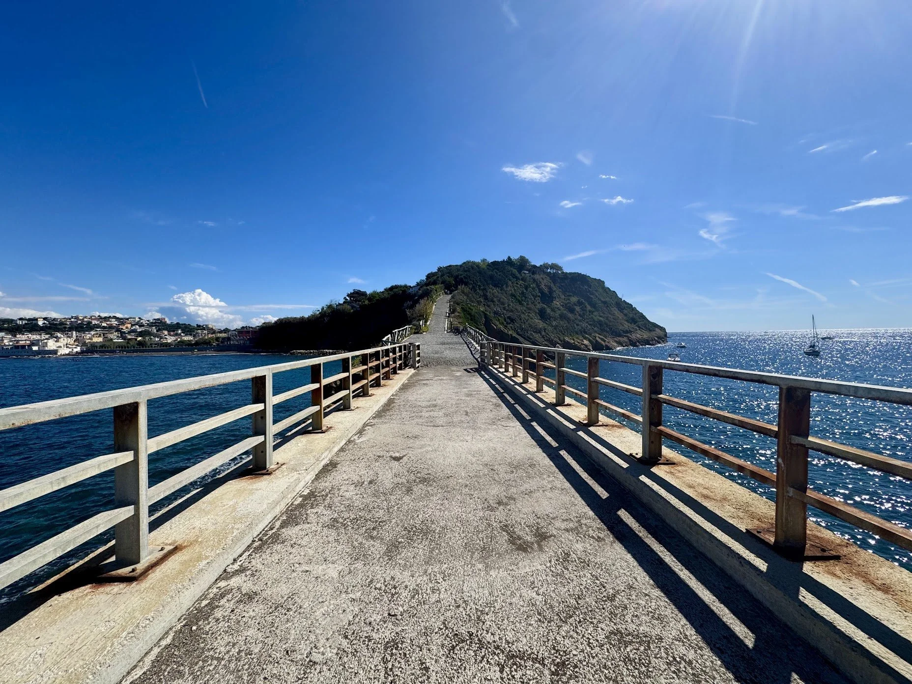 A paved walkway extends over the blue ocean, leading to a hill with greenery. Safety railings are on both sides of the walkway. The sky is clear with a few clouds and the sun is shining brightly.