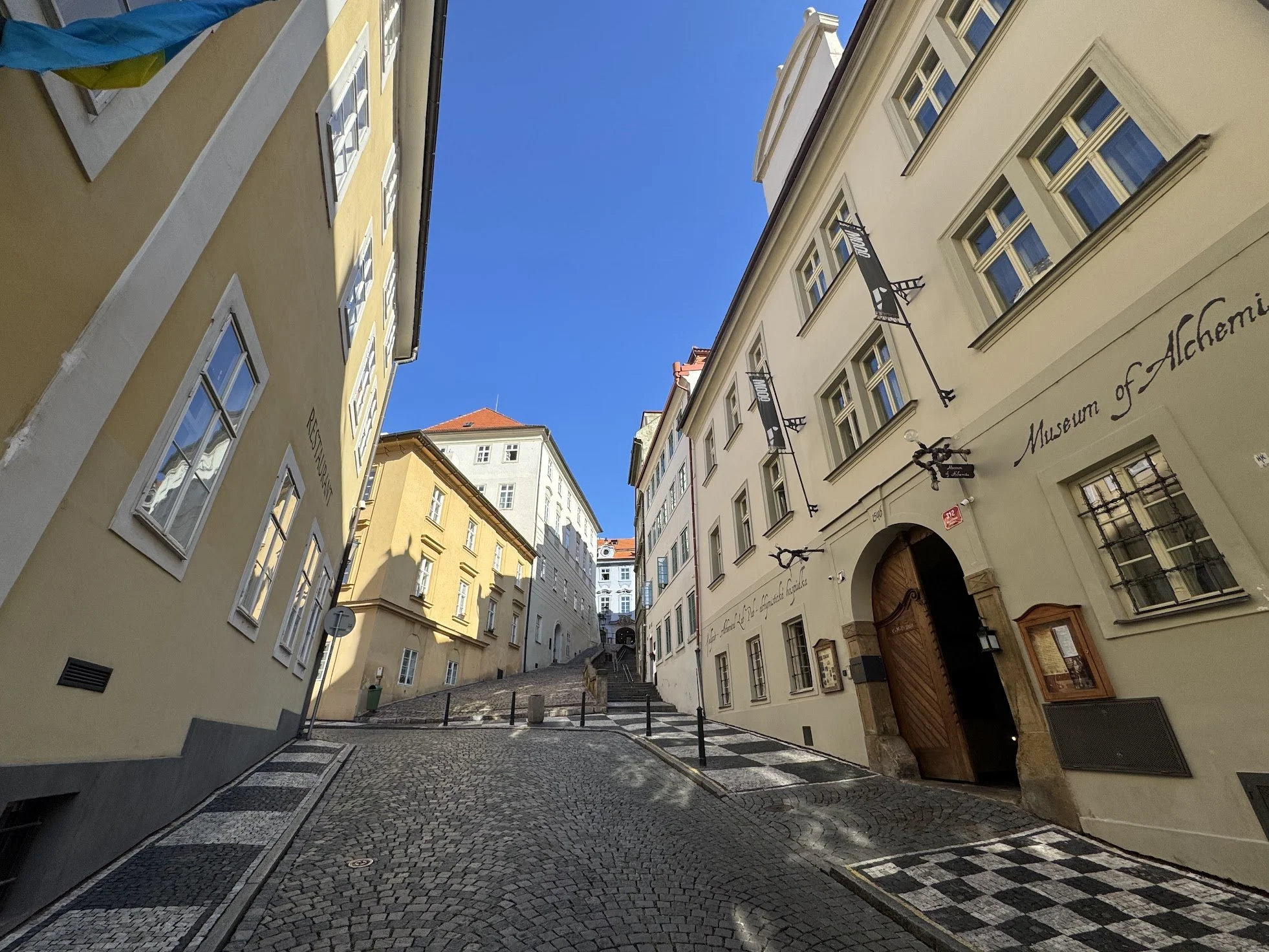 A narrow cobblestone street in a European city, lined with historic buildings, some with white and yellow facades, and signs indicating museums and shops. The sky is clear and blue.