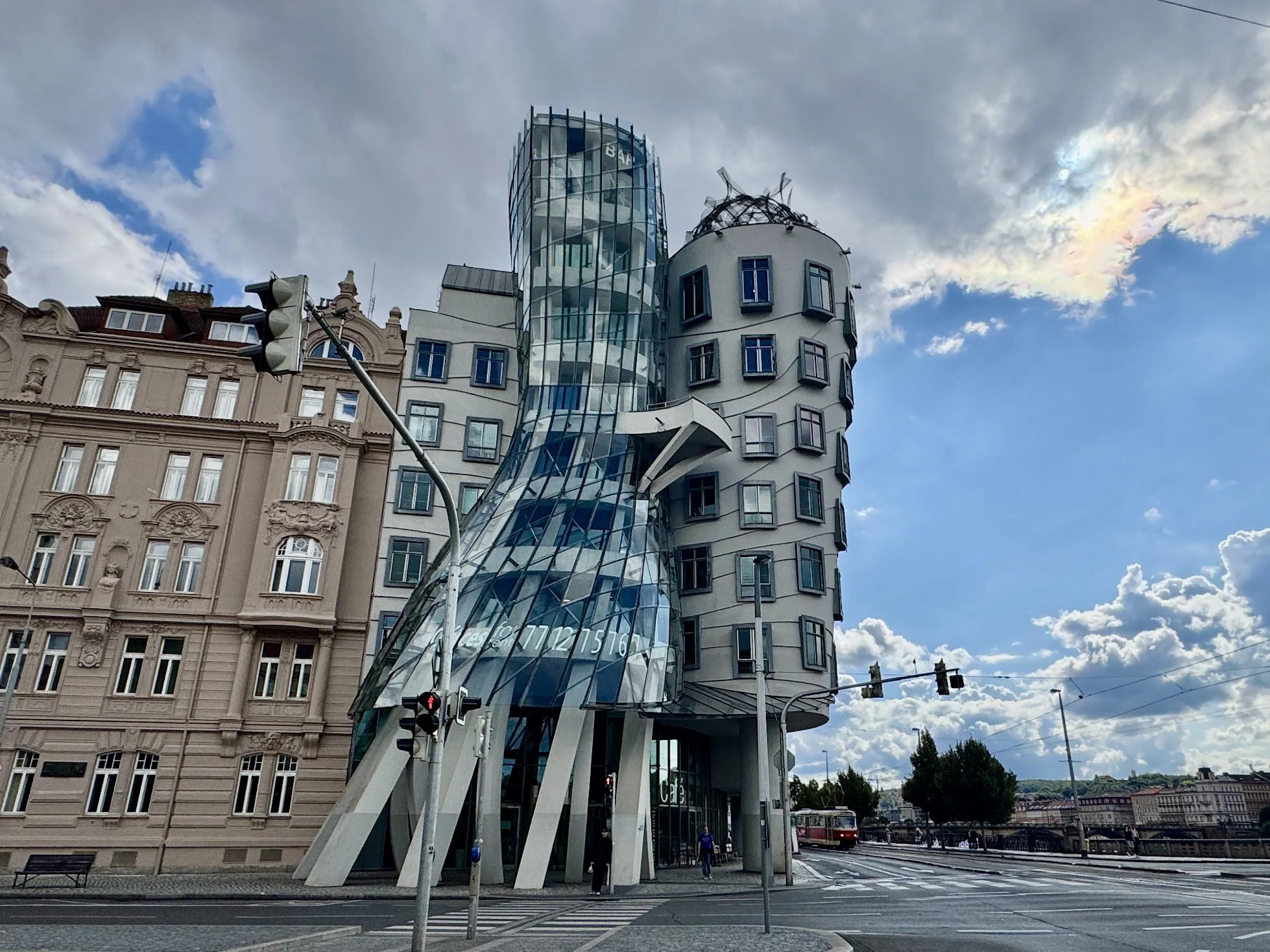 Modern architectural building with a glass spiral staircase and irregularly shaped windows, located next to traditional historic buildings, with street traffic and pedestrians in the foreground under a partly cloudy sky.