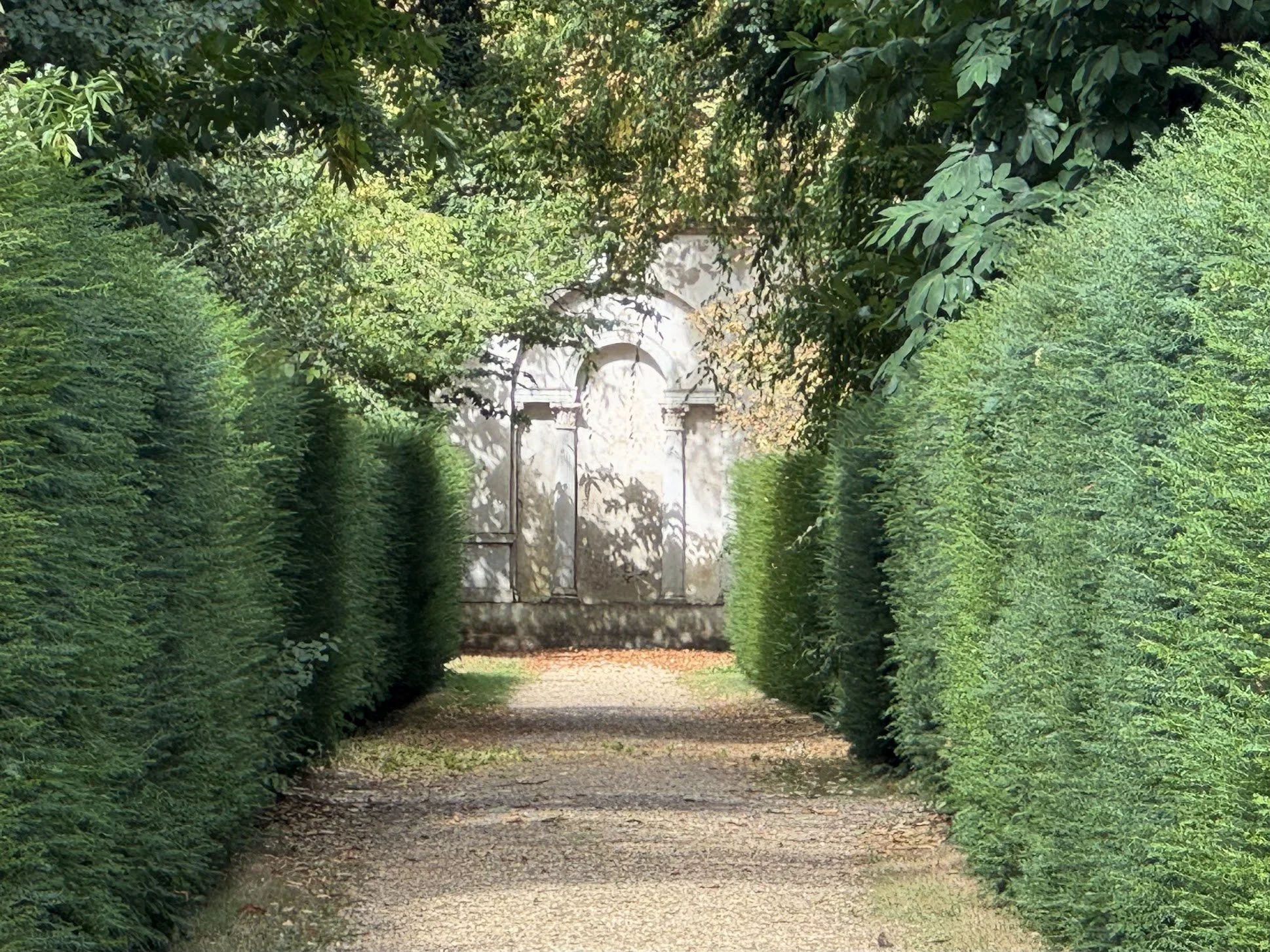 A gravel pathway surrounded by lush green hedges on both sides, leading to a stone arch gateway in the distance.