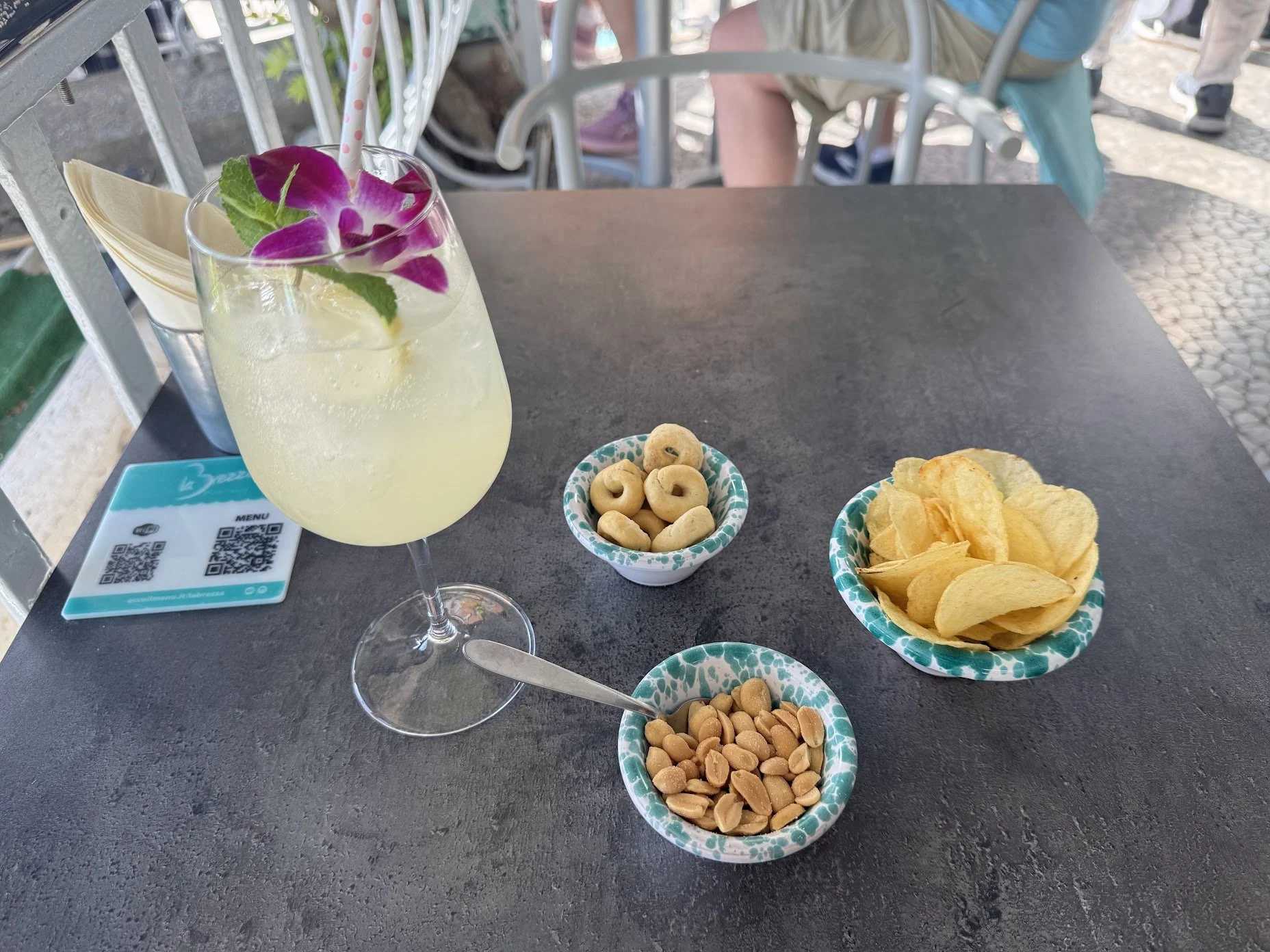 A table setting with a glass of yellow cocktail garnished with purple and green edible flowers, with small bowls of snacks such as potato chips, salted peanuts, and roasted chickpeas.