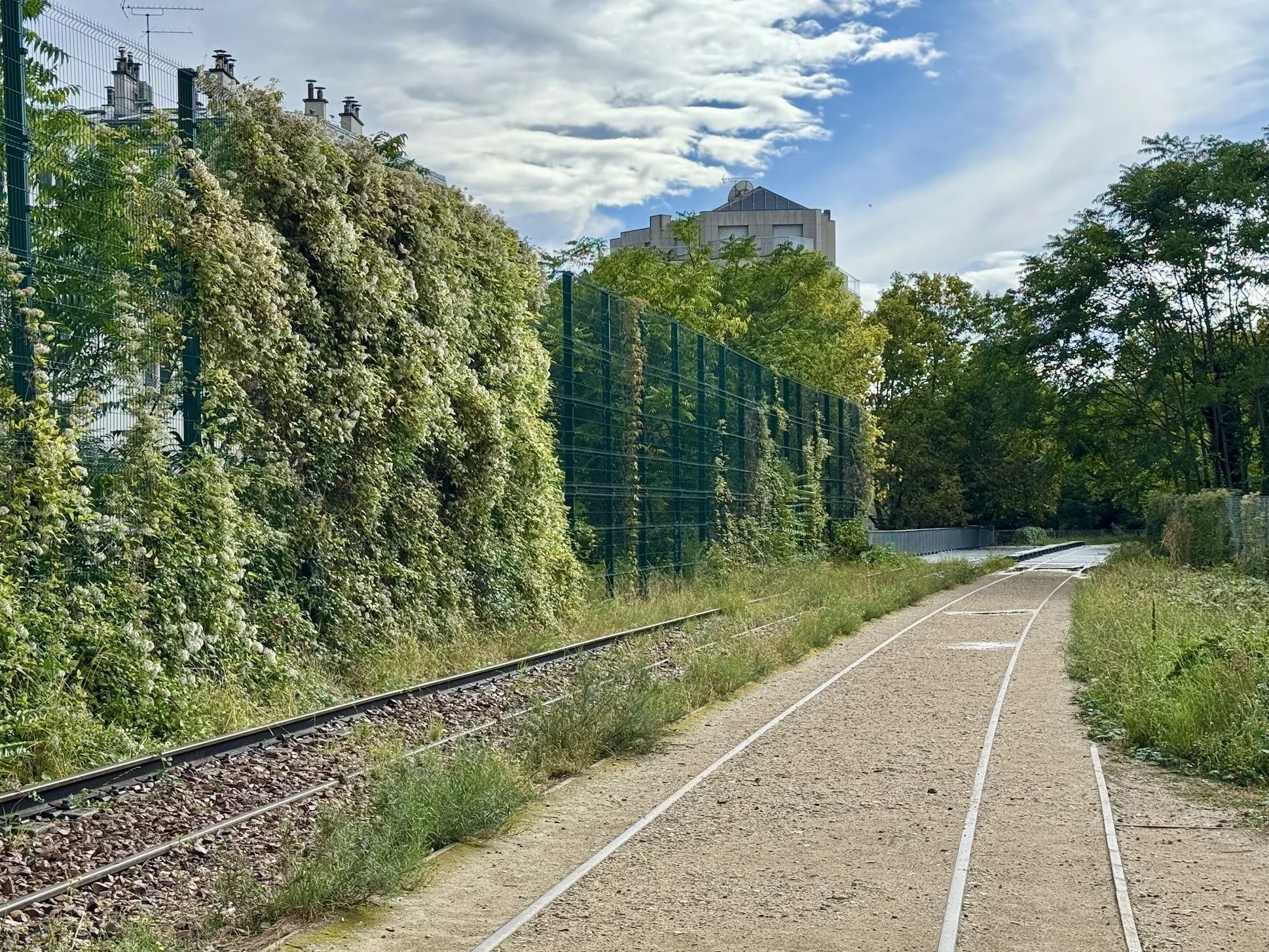 A landscape with a railway track surrounded by green trees and a fenced wall on one side, with a modern building visible in the background under a partly cloudy sky.