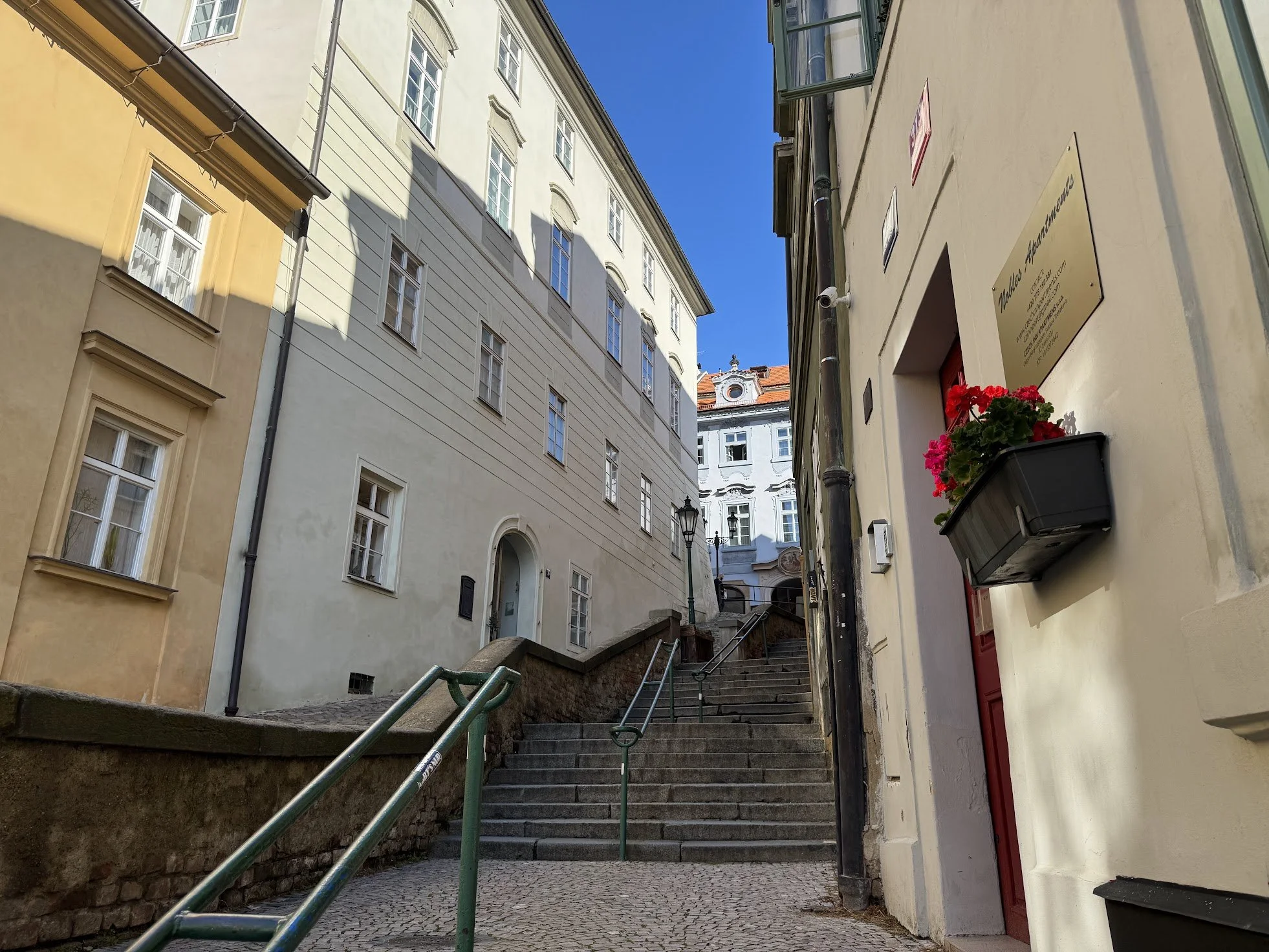 A narrow street with stone stairs and green railings leading uphill, surrounded by light-colored buildings with multiple windows. A sign and a flower box are visible on the right side, and the sky is clear and blue.