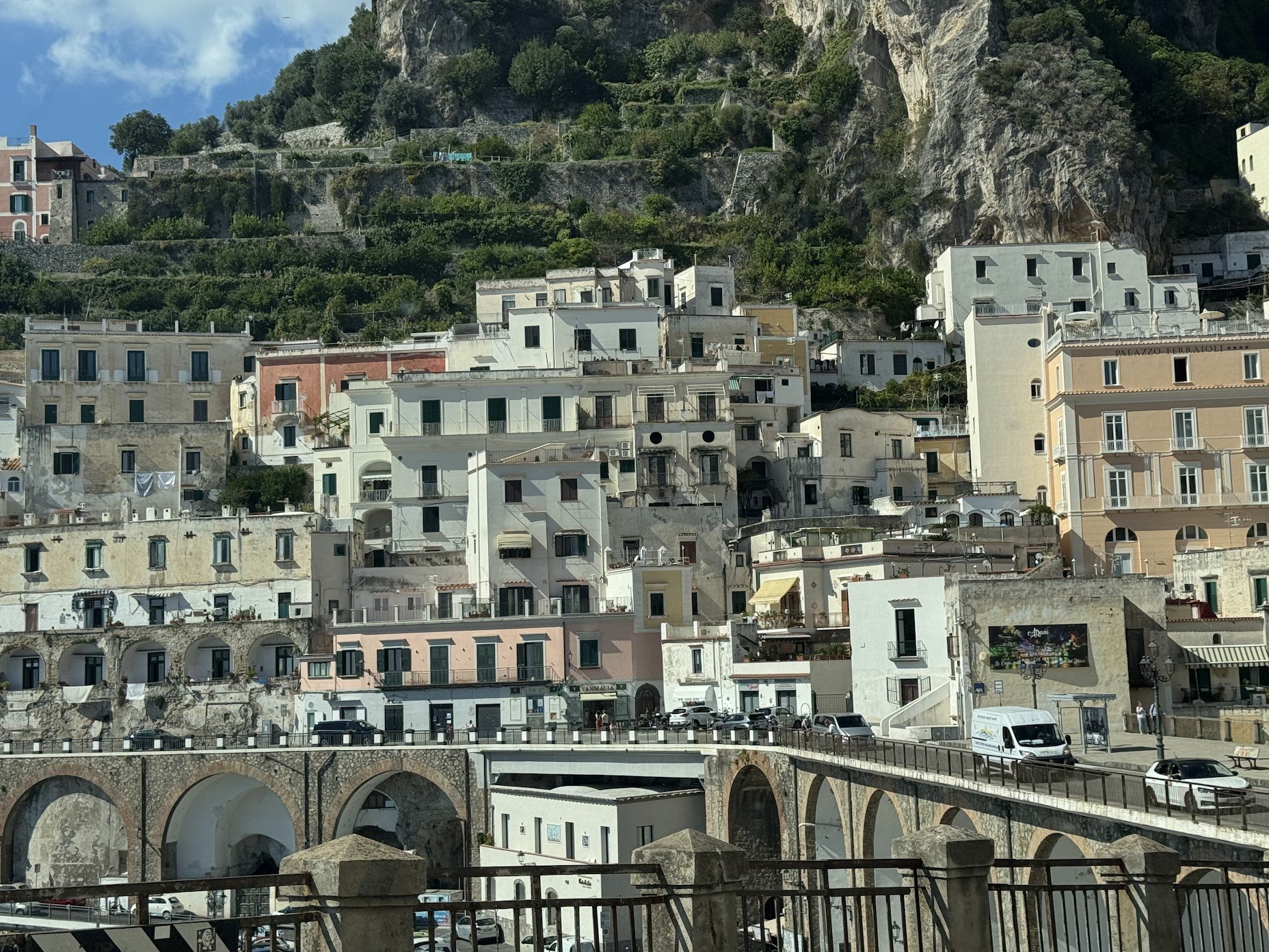Colorful hillside buildings with white, beige, and pink facades, many with balconies, terraced into a steep hillside with greenery, trees, and rocky cliffs in the background, and a stone bridge in the foreground with cars and people.