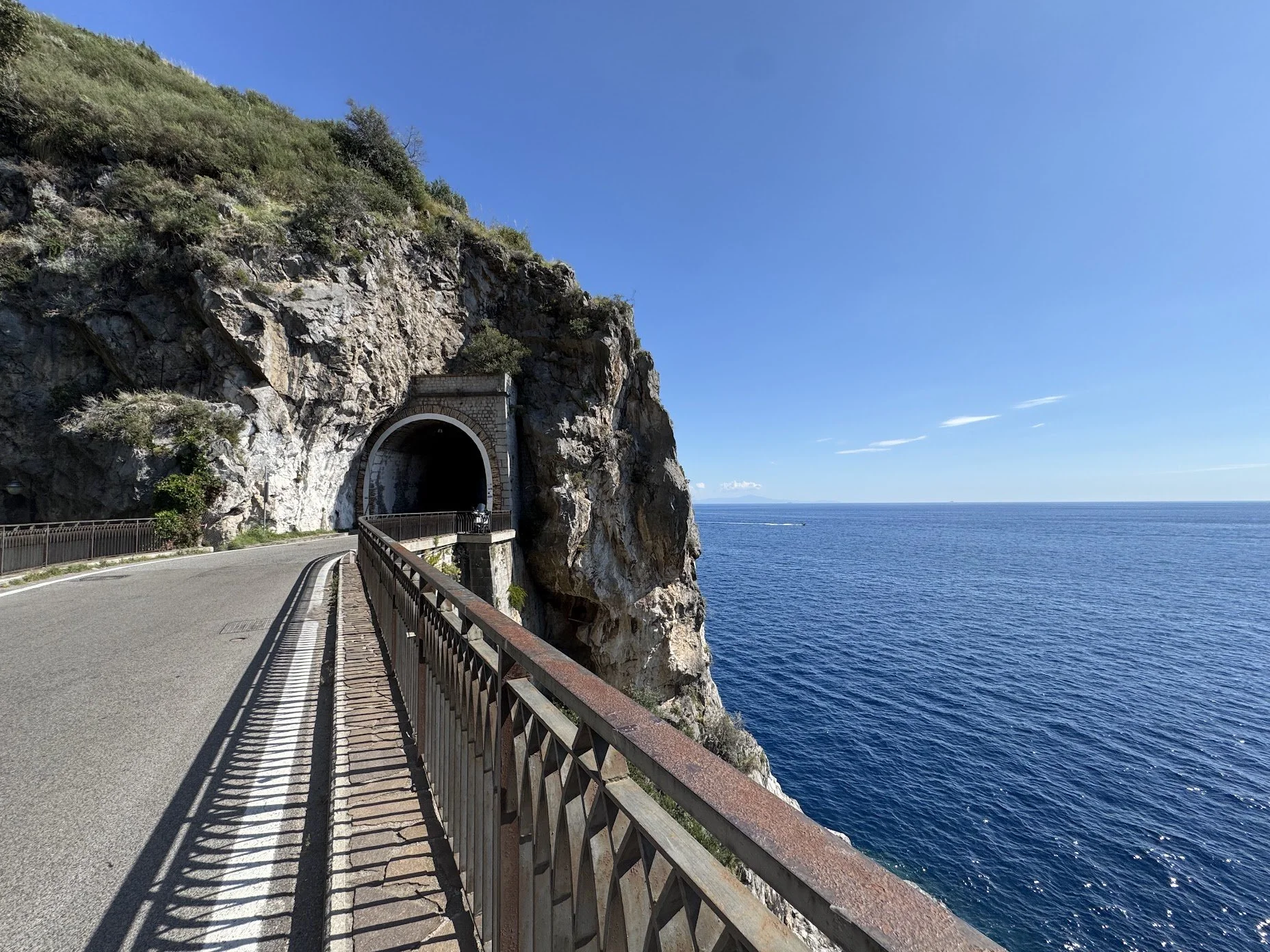 A coastal road carved into a rocky cliffside overlooking the ocean, with a tunnel entrance in the cliff face and a motorcycle on the road.