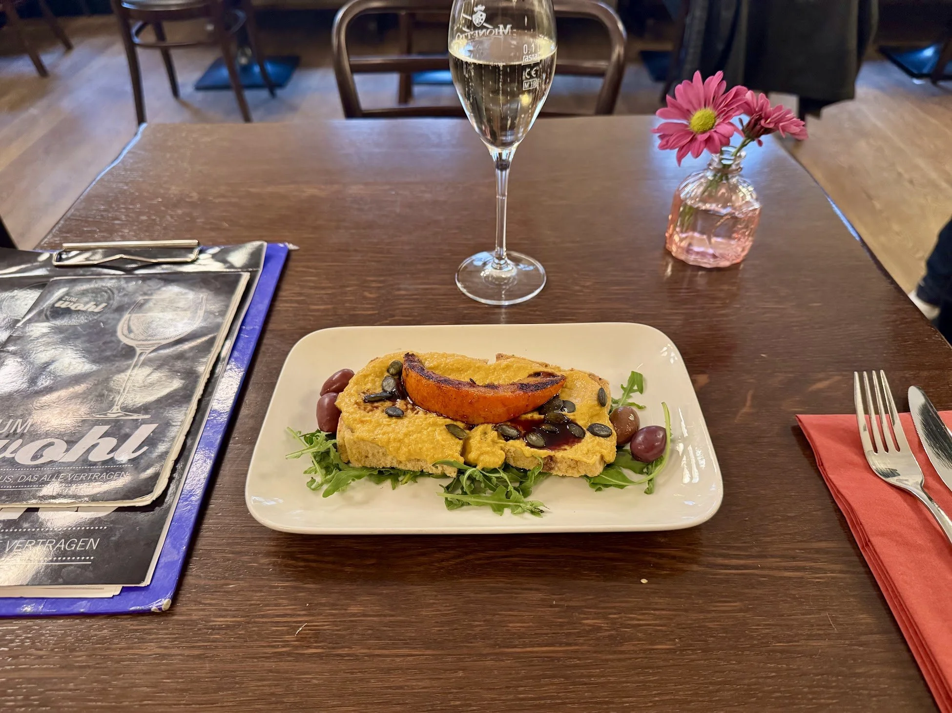 A plated slice of pumpkin pie garnished with pumpkin seeds, a cherry tomato, and some greens on a white rectangular plate, with a glass of champagne, a pink flower in a glass vase, and a black and blue menu on a wooden table.