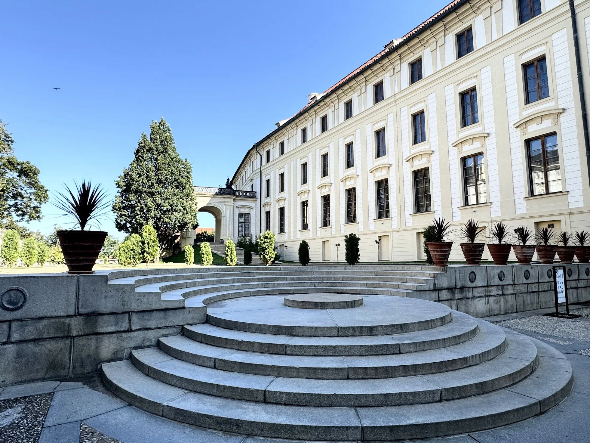 A historical white building with large windows and a red tiled roof. In the foreground, there are stone steps leading up to the building, flanked by potted plants and lush green trees under a clear blue sky. A single bird is flying in the sky.