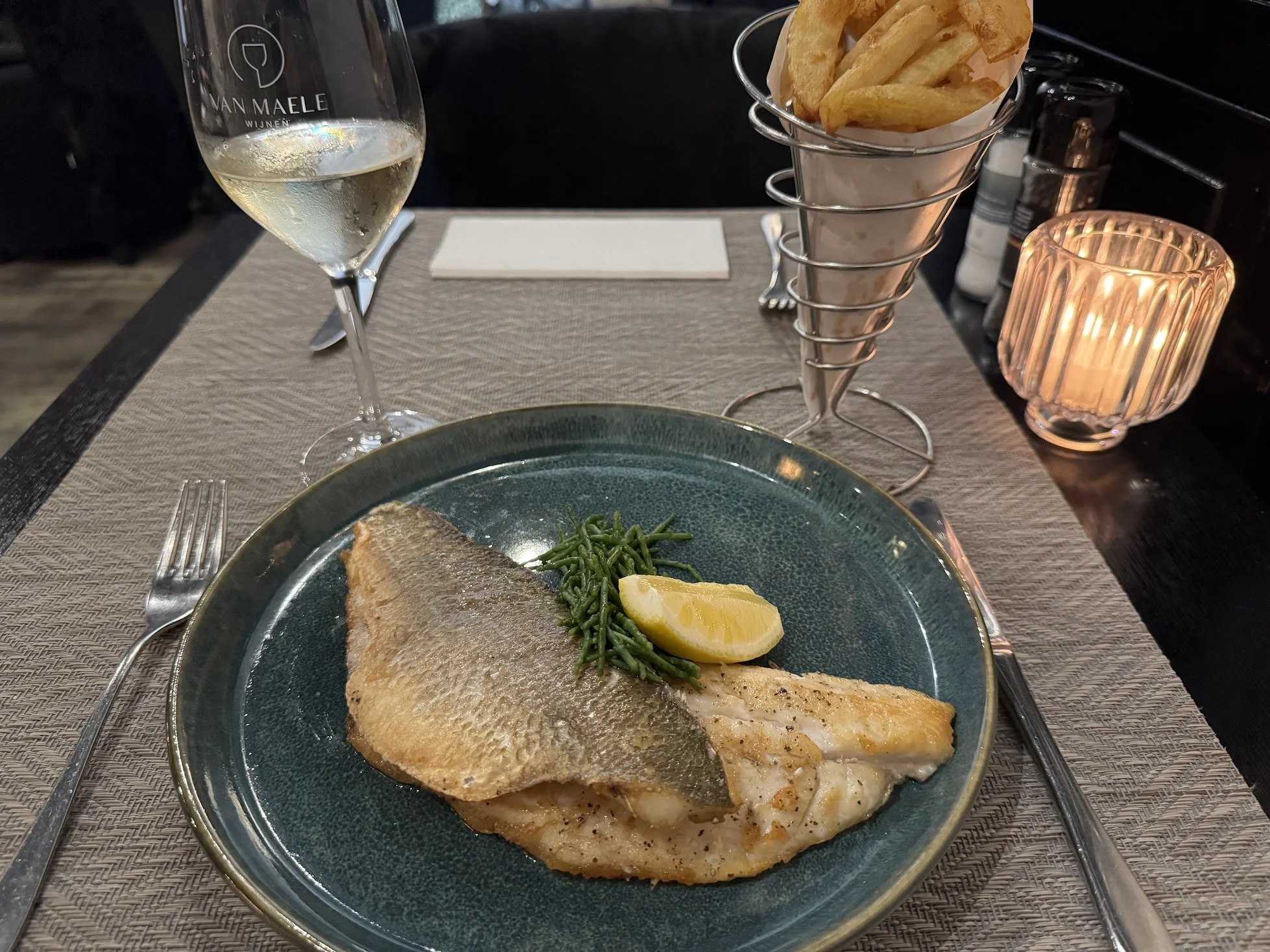 A plate with a piece of cooked fish, green beans, and a lemon wedge, on a dining table. In the background, there's a glass of white wine, a cone of French fries in a metal holder, a candle, and some salt and pepper shakers.