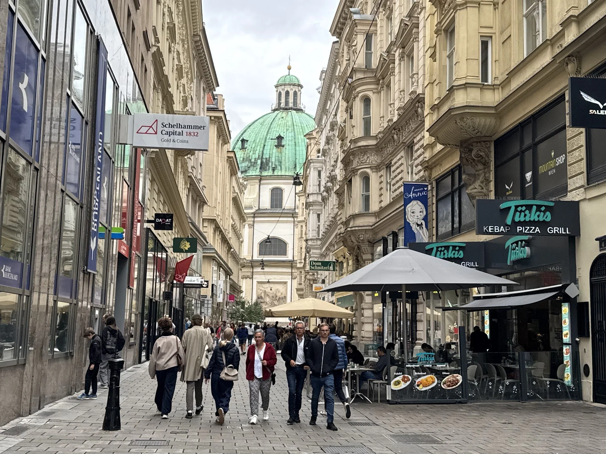 A busy pedestrian street in a European city with historical buildings on both sides and a domed religious building with a green roof in the background. There are commercial signs and outdoor seating from cafes and restaurants, with people walking and