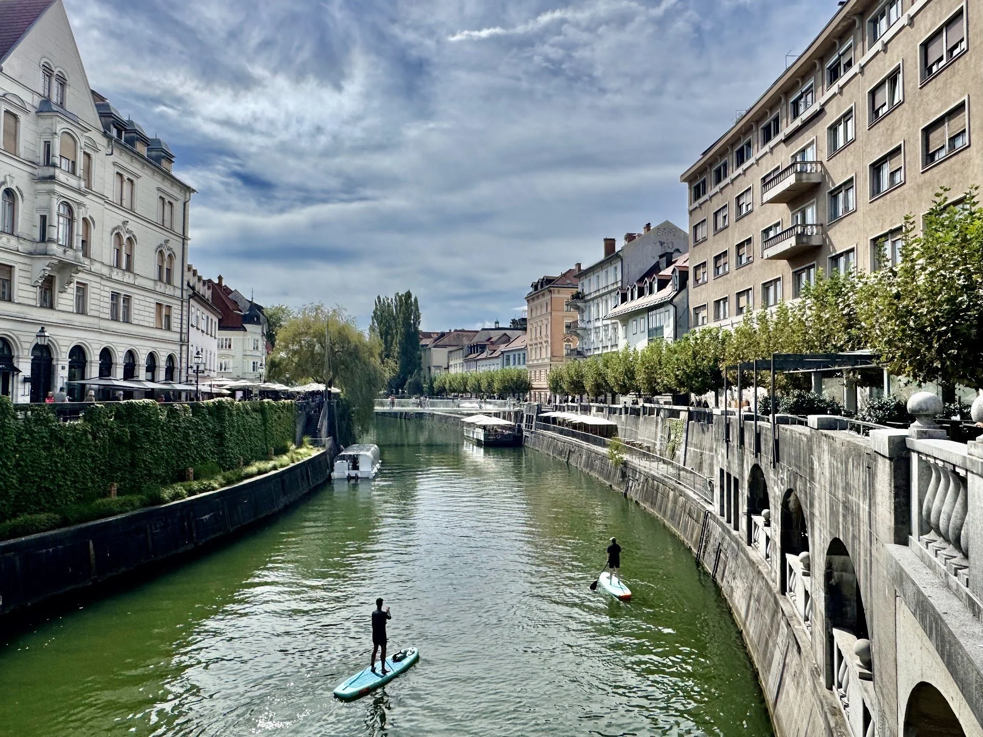 Two people stand on paddleboards on a canal, with buildings on each side and a cloudy sky above.