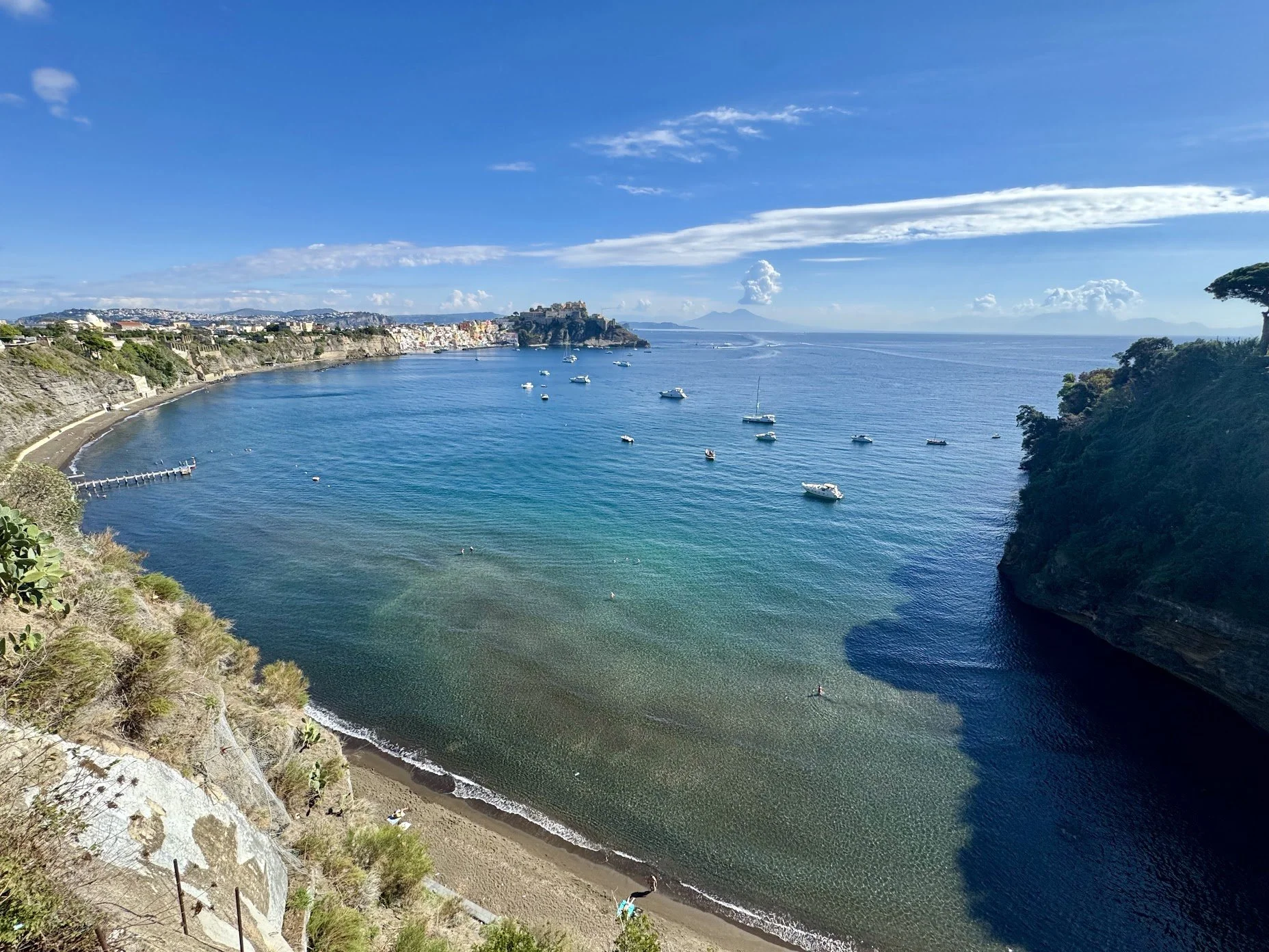 Coastal view of a bay with boats floating on the water, a sandy beach with a few people, rocky cliffs covered in greenery, and a cityscape in the distance under a blue sky with scattered clouds.