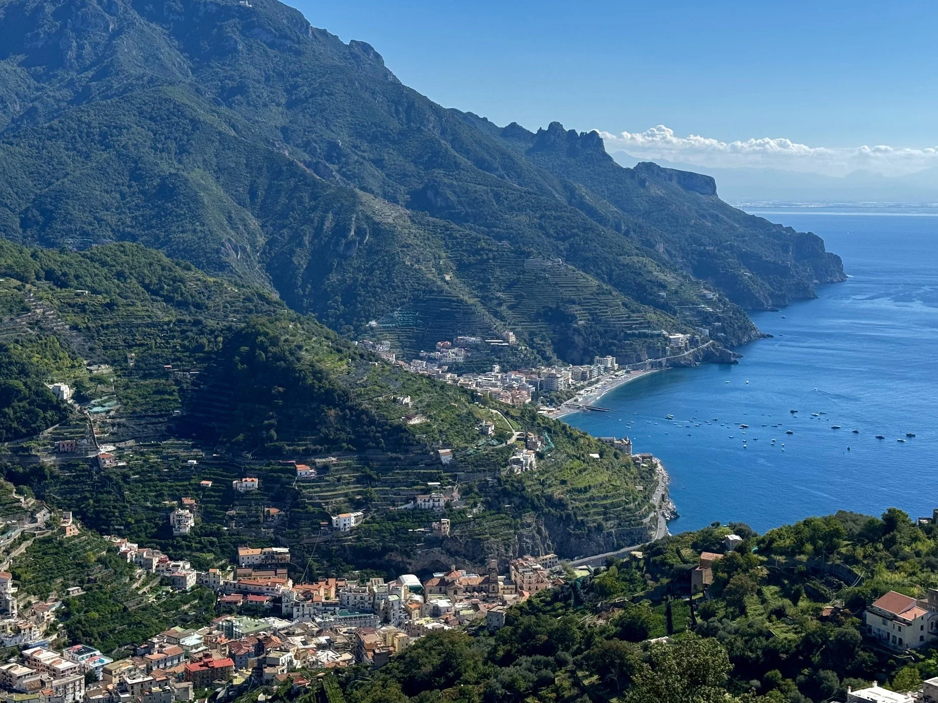 Coastal village with houses and terraced agriculture along steep hillsides, overlooking a blue sea under a clear sky with some clouds.