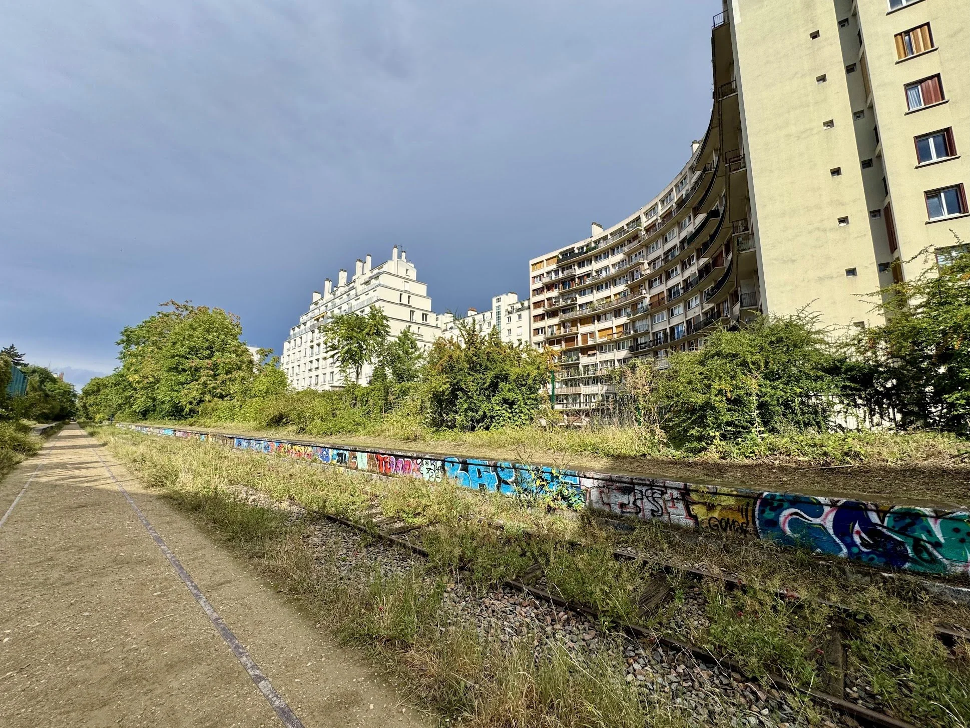 A view of tall apartment buildings in an urban area with trees and greenery in the foreground. A railroad track runs along a graffiti-covered barrier in front of the buildings. The sky is partly cloudy.