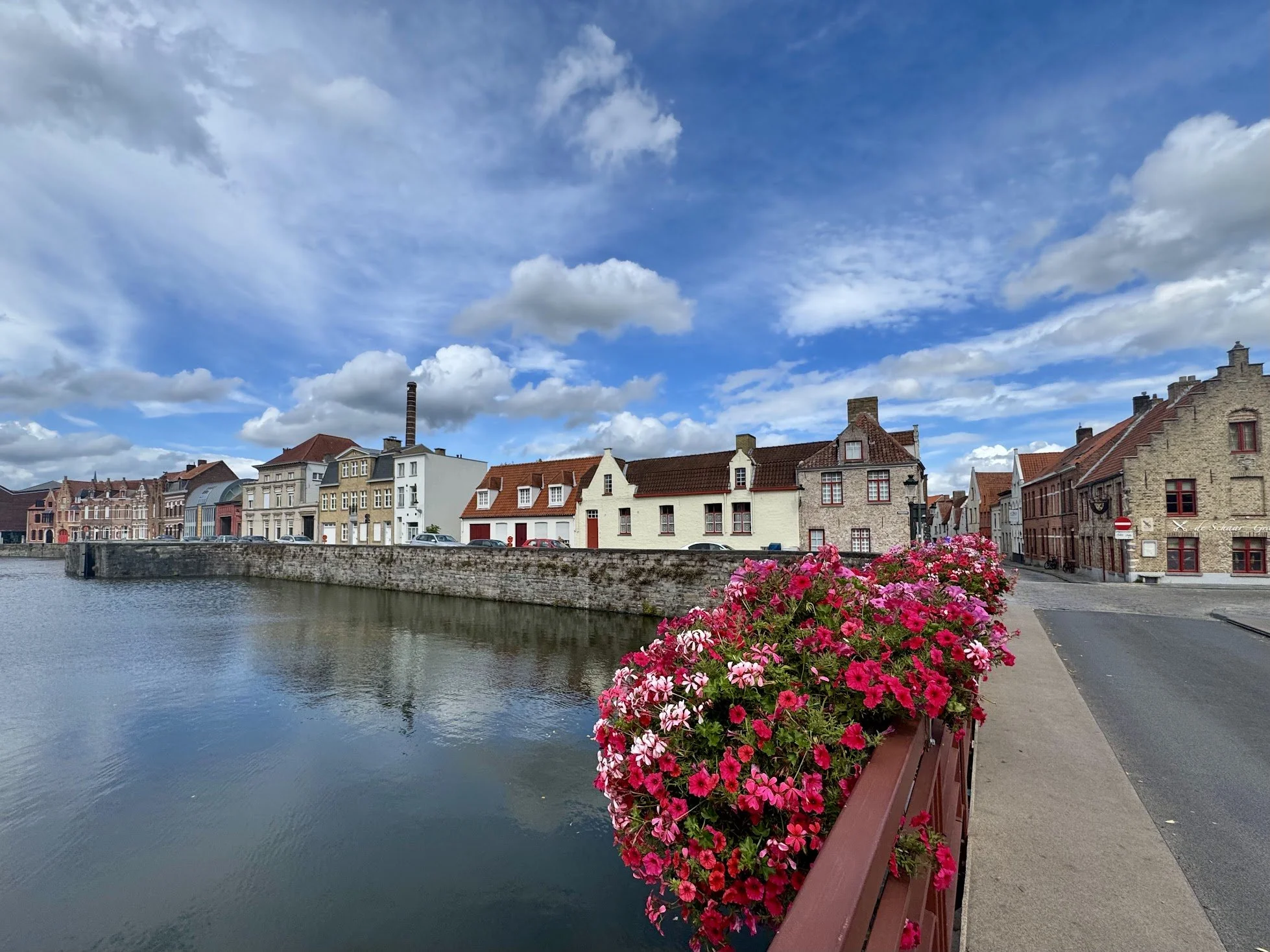 A scenic view of a canal lined with historic buildings and a flower box with pink and red flowers in the foreground, under a partly cloudy sky.