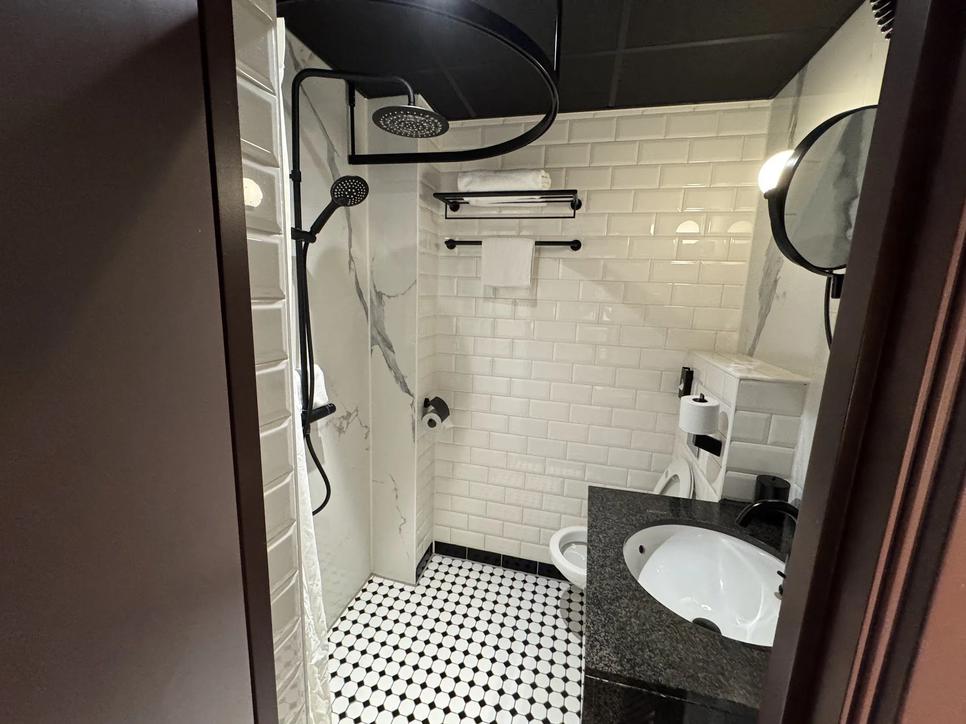 A small bathroom with white subway tile walls, a black countertop with a white sink, a toilet with the lid up, a black shower with a curved rain shower head and handheld shower head, a black towel rack with a white towel, a mirror with black framing,