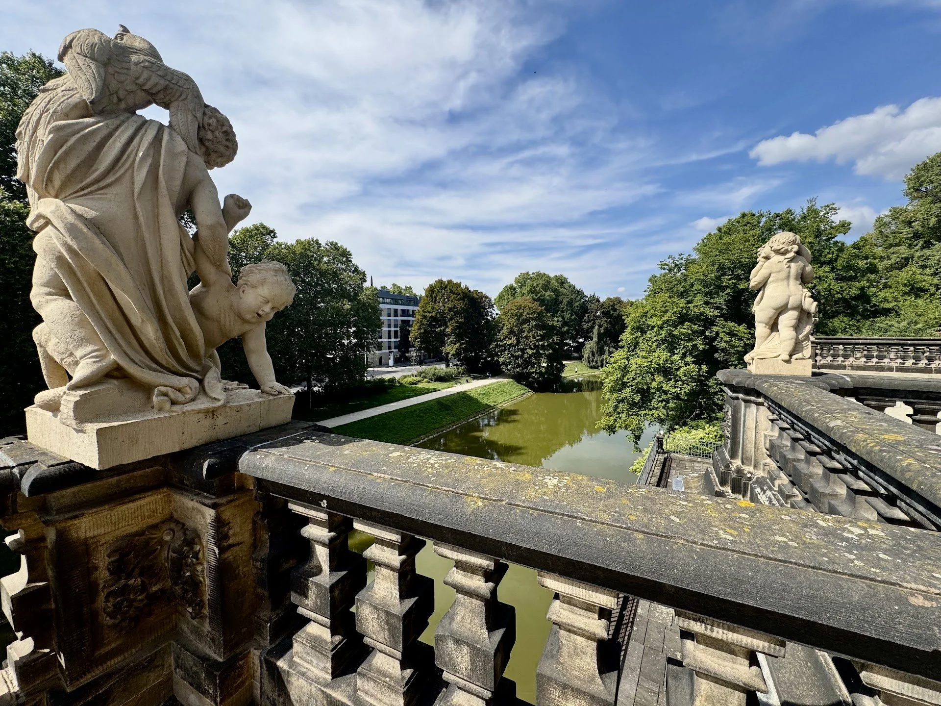 View of a stone bridge with detailed sculptures of cherubs and mythical creatures, overlooking a river with green trees and a modern building in the background under a partly cloudy sky.