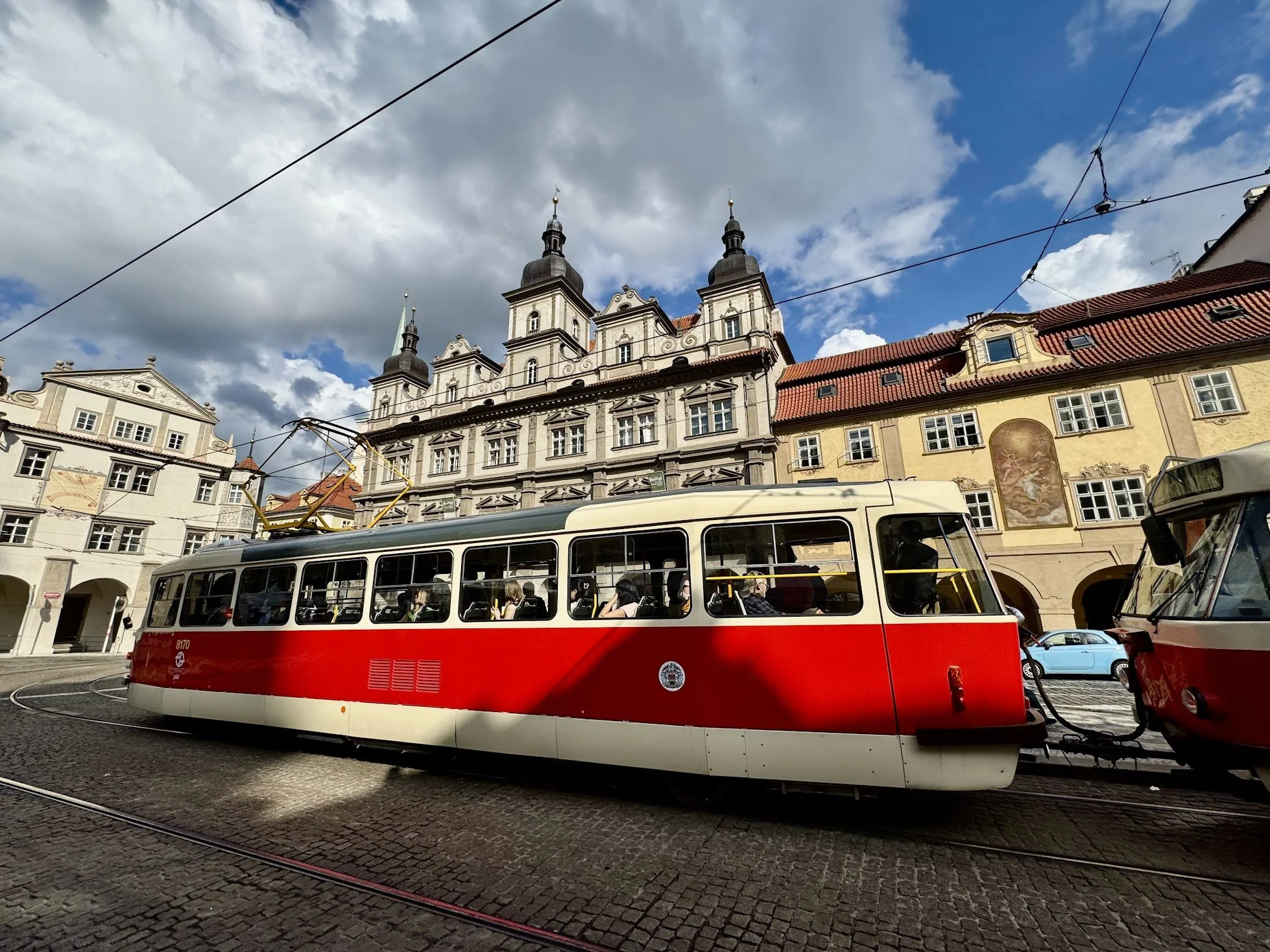 A red and white tram is moving through a European city square, with buildings featuring ornate architecture and red-tiled roofs in the background. People are on the tram, and overhead tram power lines are visible against a partly cloudy sky.