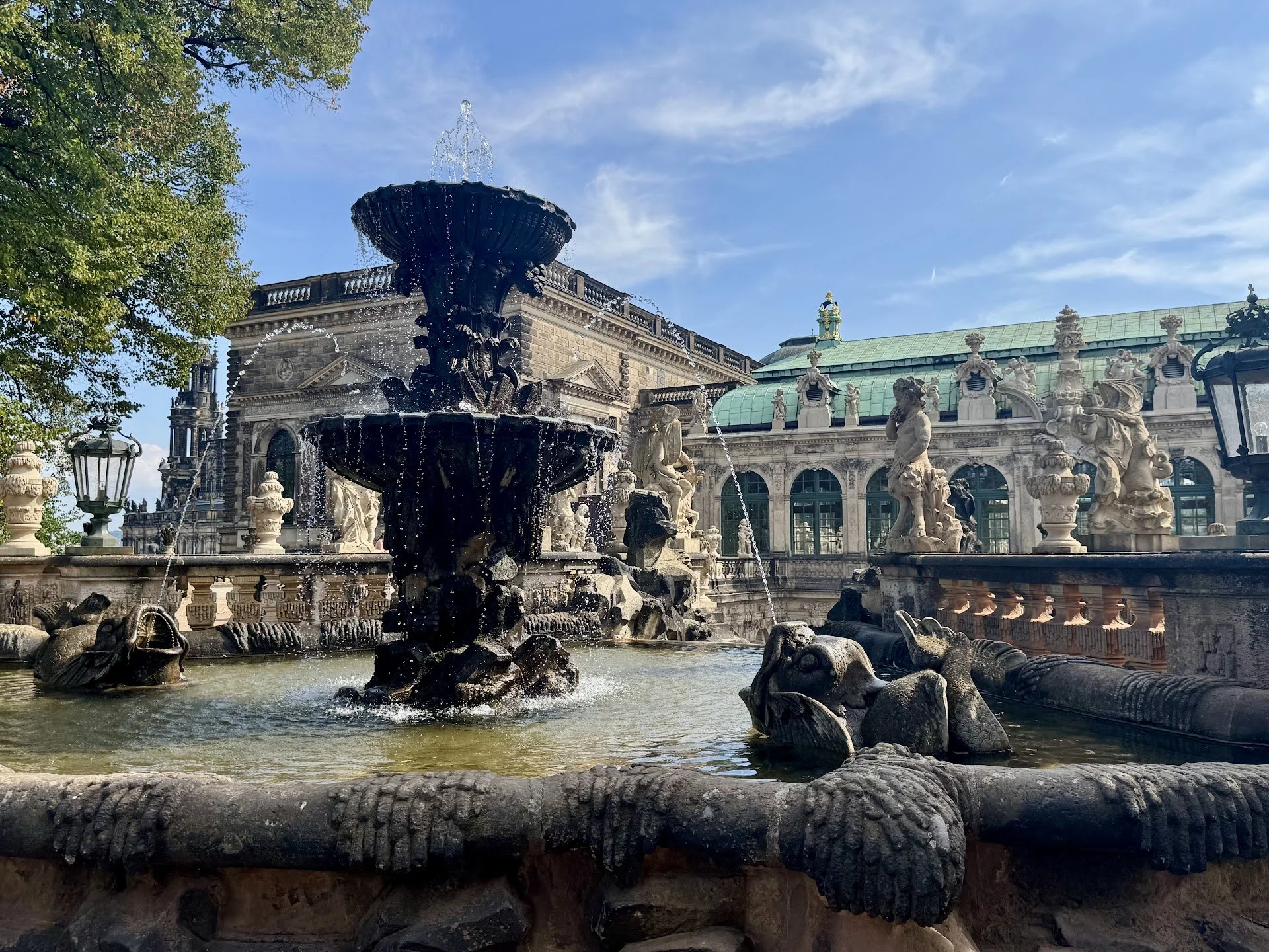 A decorative fountain with a black basin at the top, water flowing from it into the lower pool, surrounded by ornate stone sculptures of fish and mythical creatures, with a historic building featuring statues on its roof and large windows in the back
