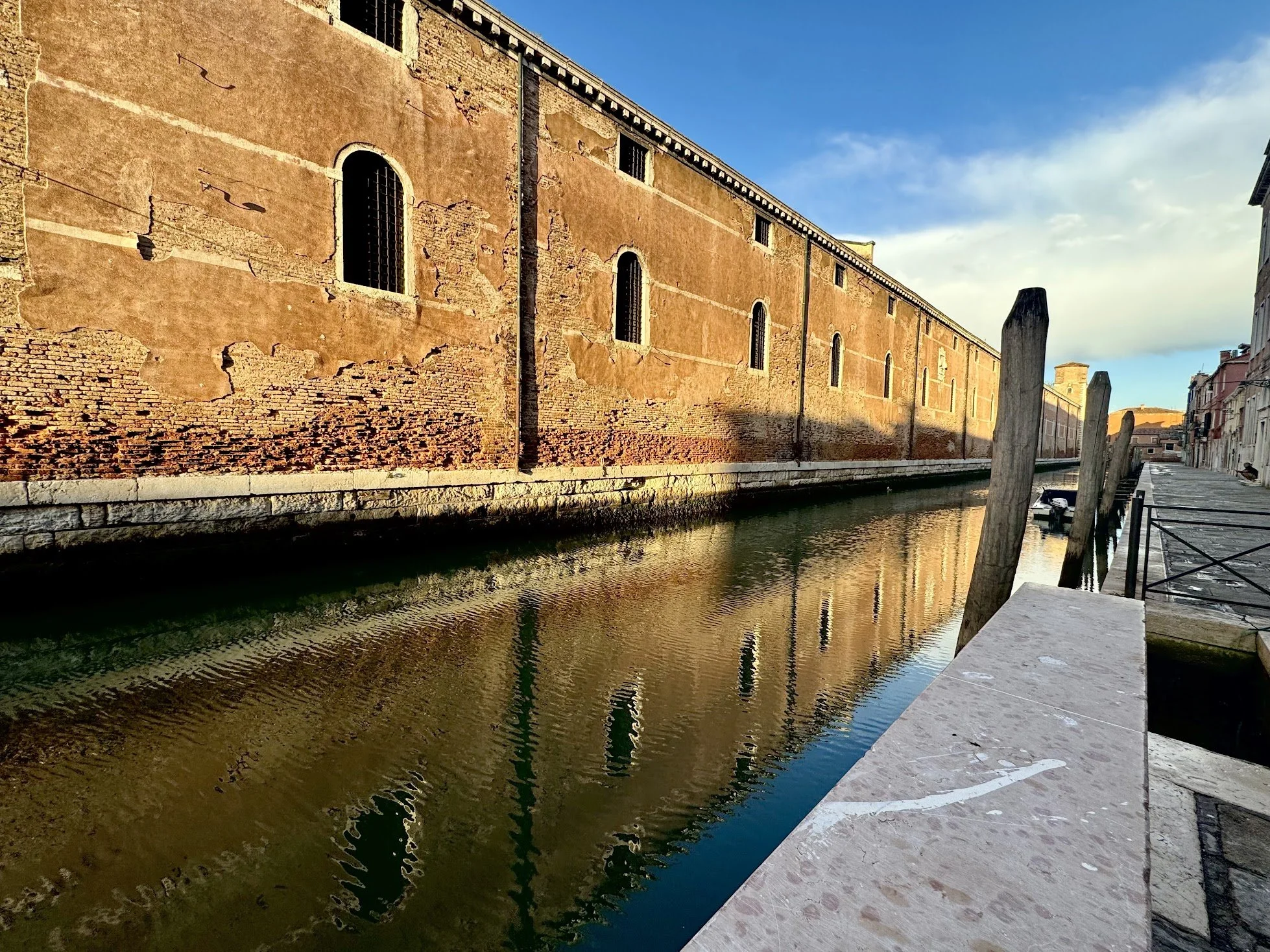 A weathered brick and plaster building along a canal with water and a stone walkway in Venice, Italy, under a partly cloudy sky