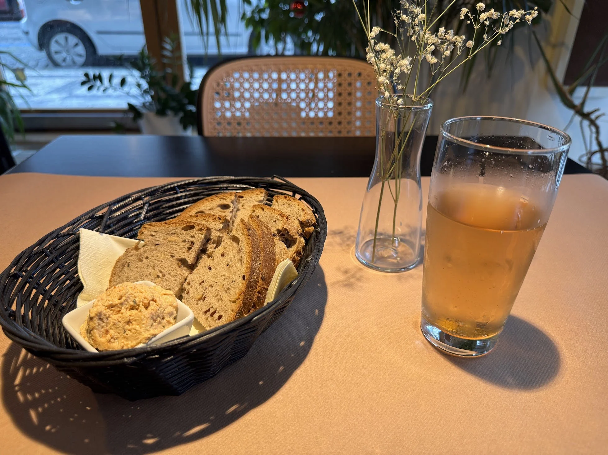 A table set with slices of fruit and nut bread in a black basket, a small dish of spread, a glass of iced tea, and a vase with white flowers. In the background, a window shows parked cars outside.
