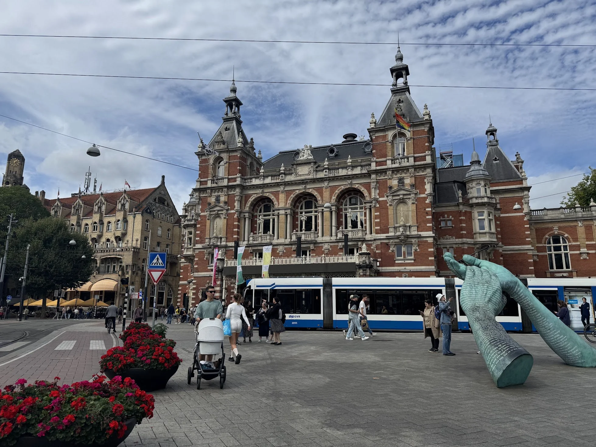 Crowd of people walking and waiting in a busy city square with historic buildings, a tram, and a large sculpture of a hand.