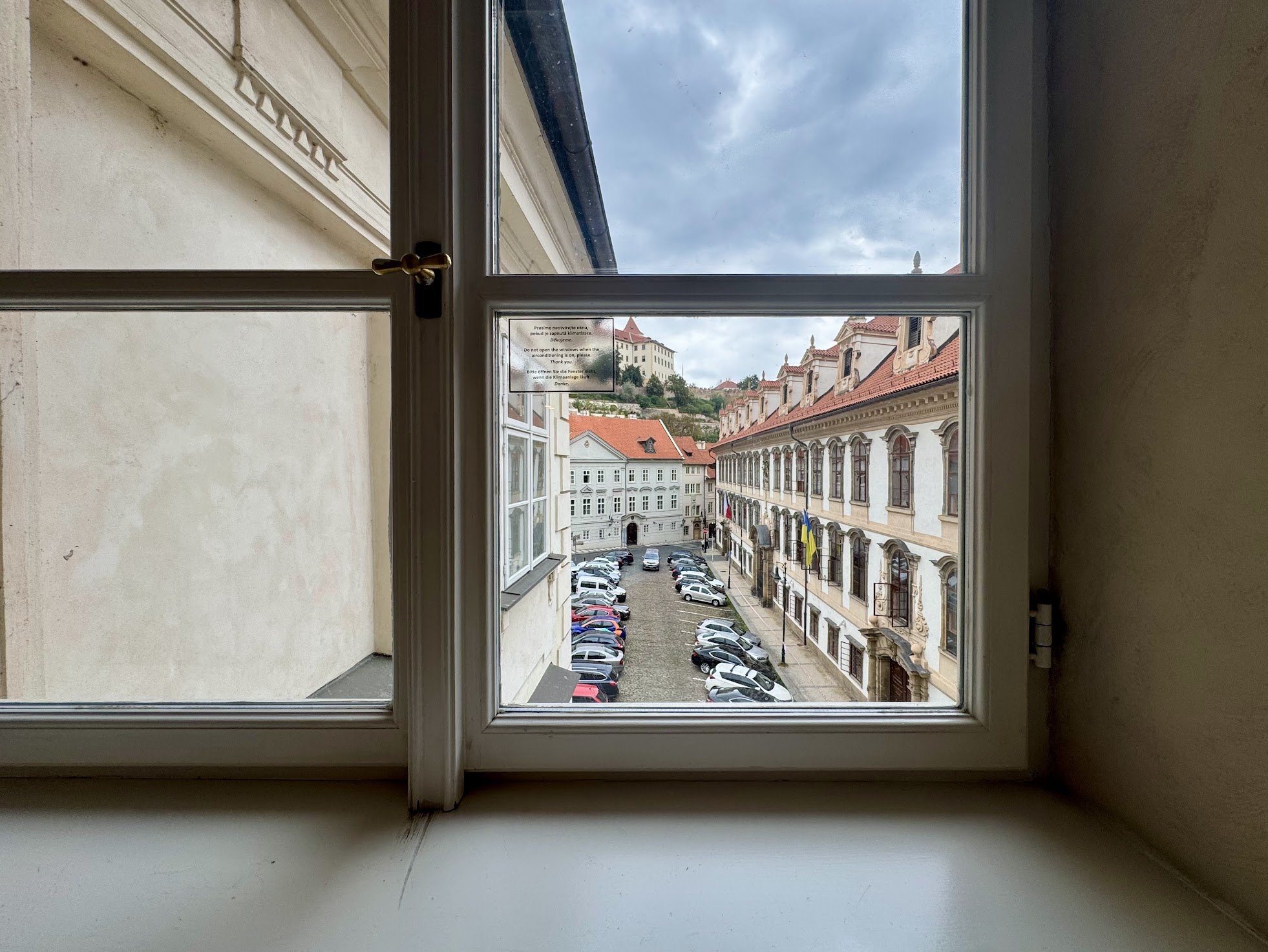 View through a window showing a street with parked cars, a historic building with ornate architecture, and a hillside with more buildings and a cloudy sky.