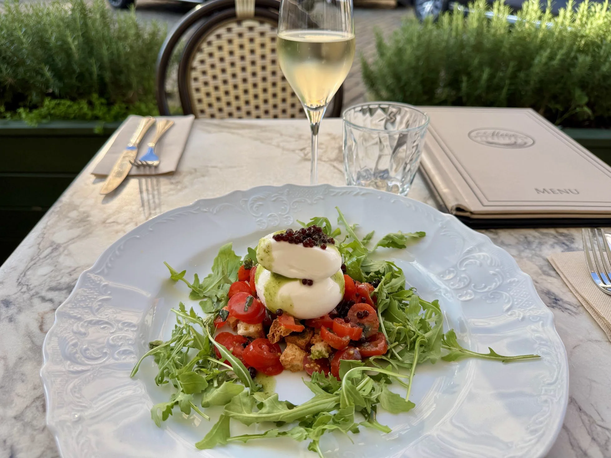 A plate of salad with greens, cherry tomatoes, avocado, and what appears to be burrata cheese topped with black caviar, on a white ornate plate. In the background, there is a glass of white wine, a glass of water, a menu, and a set of silverware on a marble table outdoors.