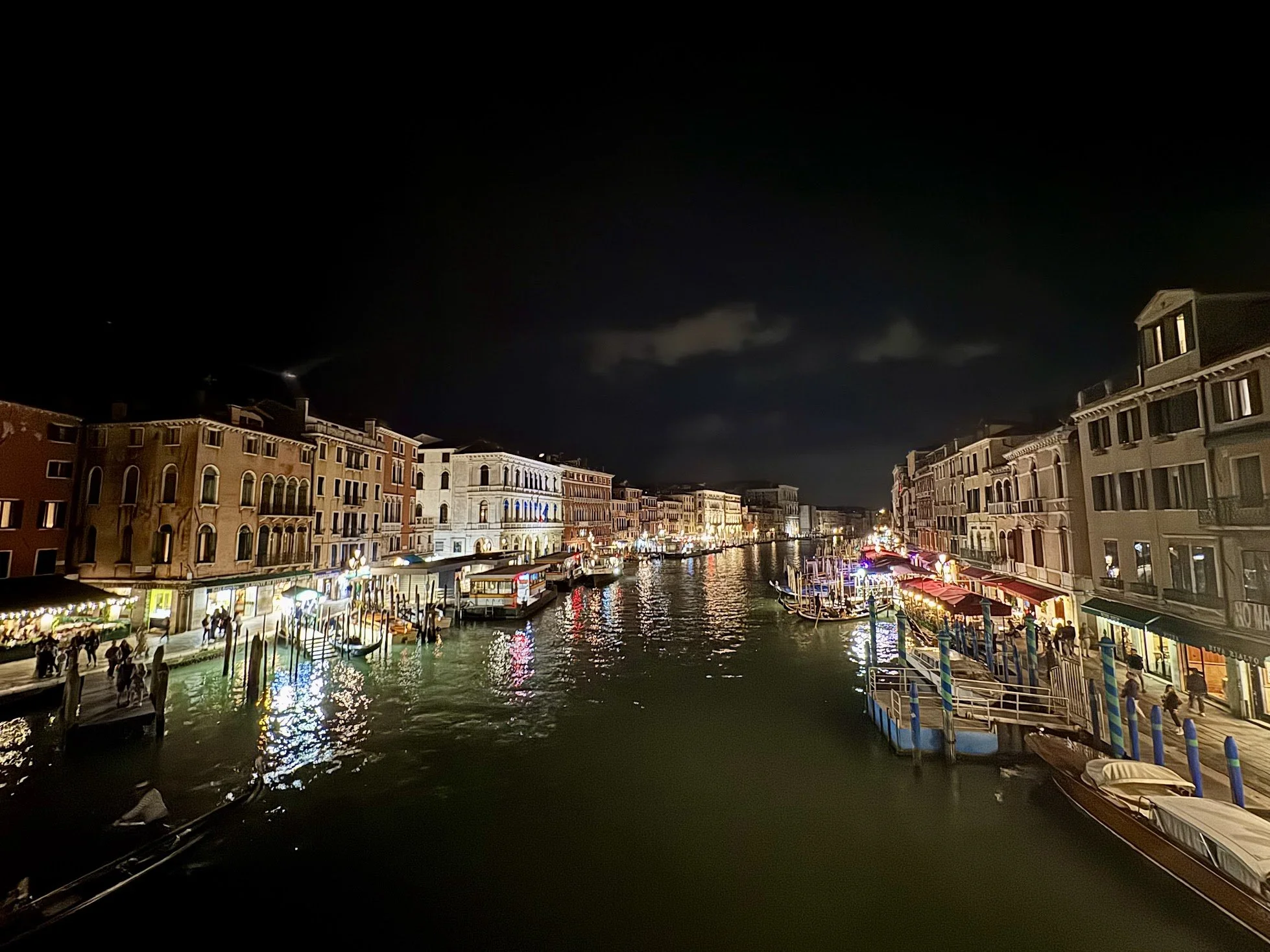 Night scene of a canal lined with colorful buildings, illuminated storefronts, and boats docked along the water, with cloudy skies overhead.