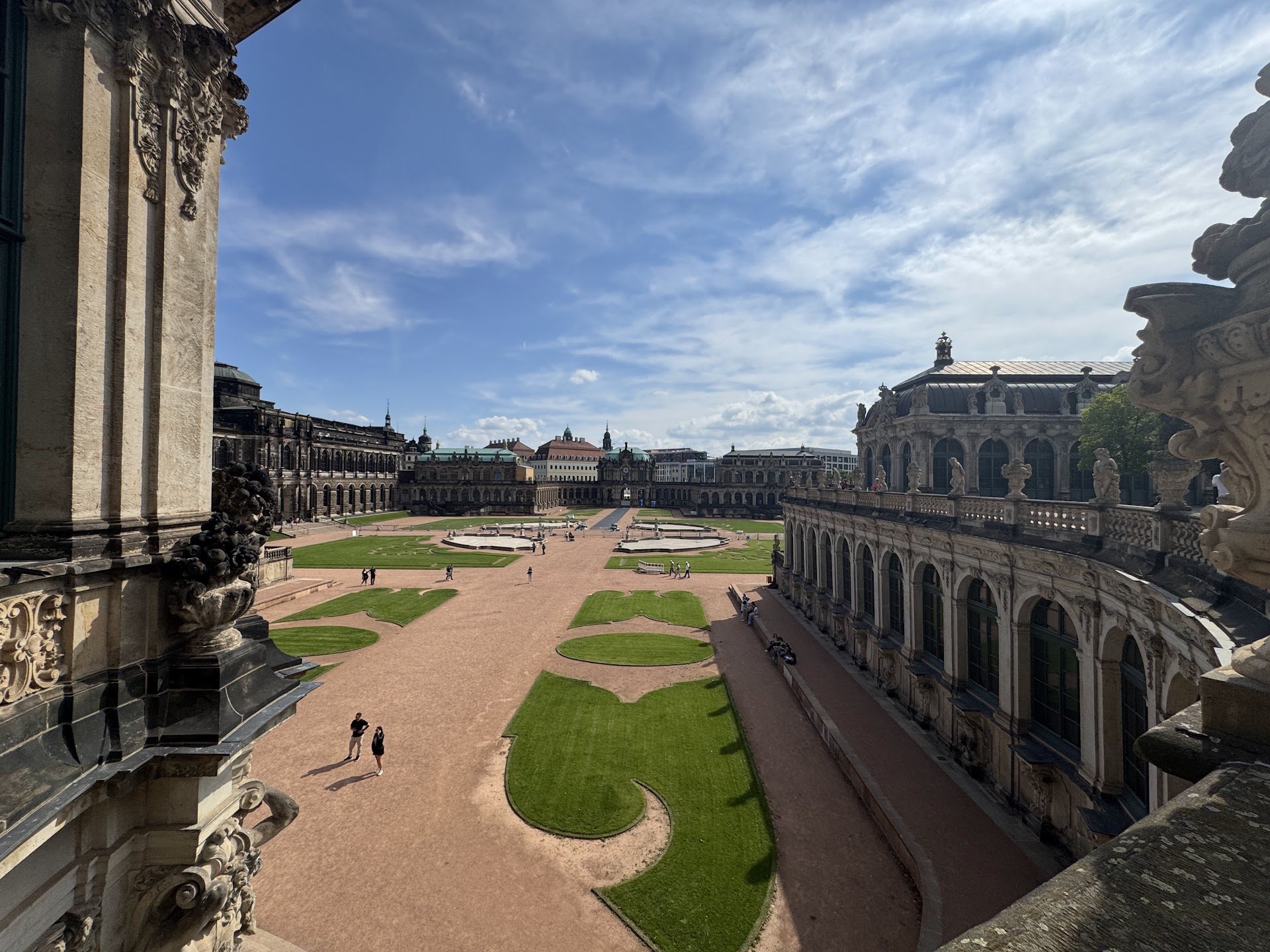View from a balcony overlooking a historic city square with ornate buildings, sculptures, and a well-maintained garden with winding pathways, under partly cloudy skies.