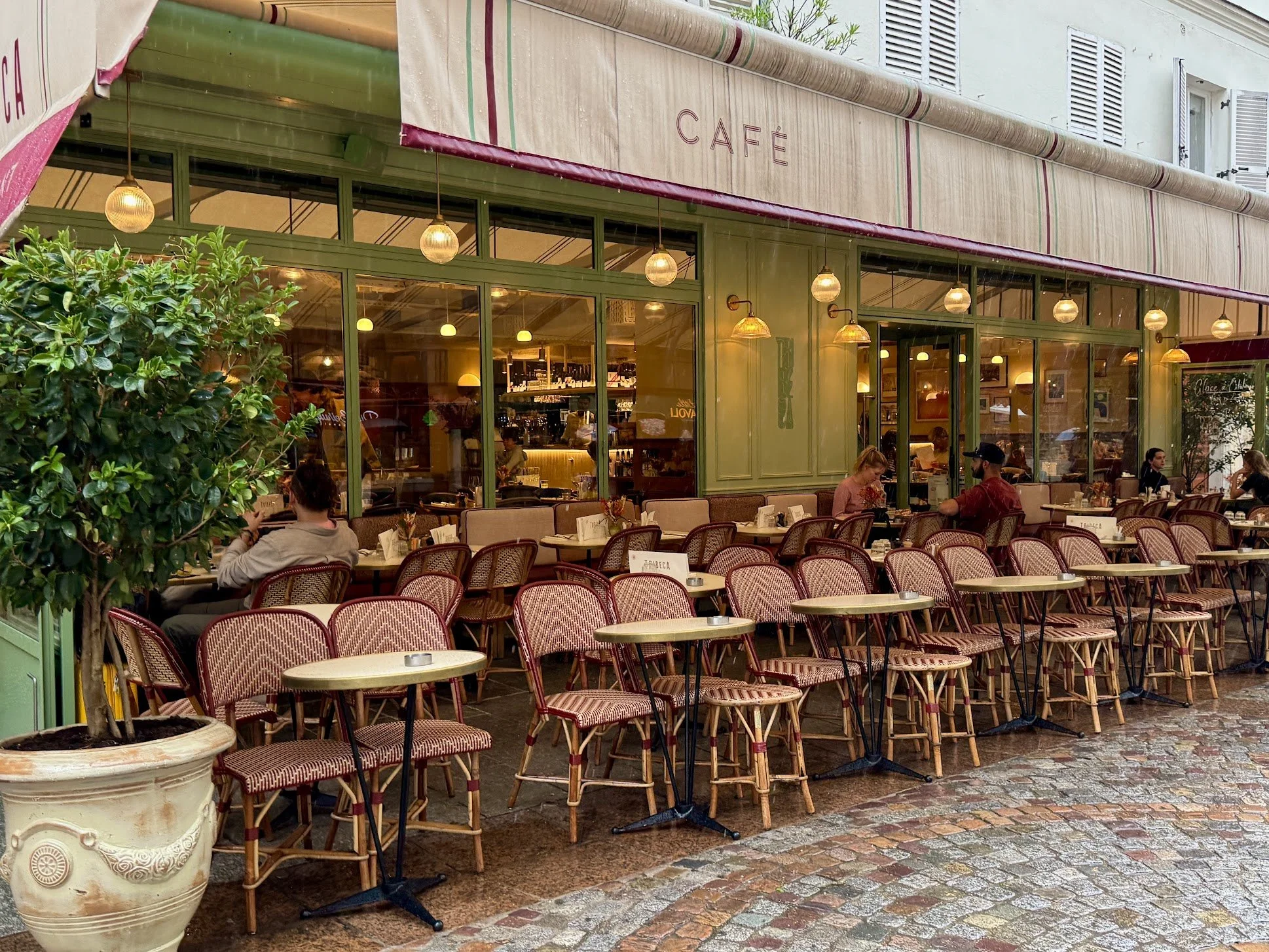 The image shows an outdoor seating area of a cafe with numerous tables and chairs, some of which are occupied by people. There are green framed windows and an entrance to the cafe, with warm lighting inside. A large potted plant is visible on the lef