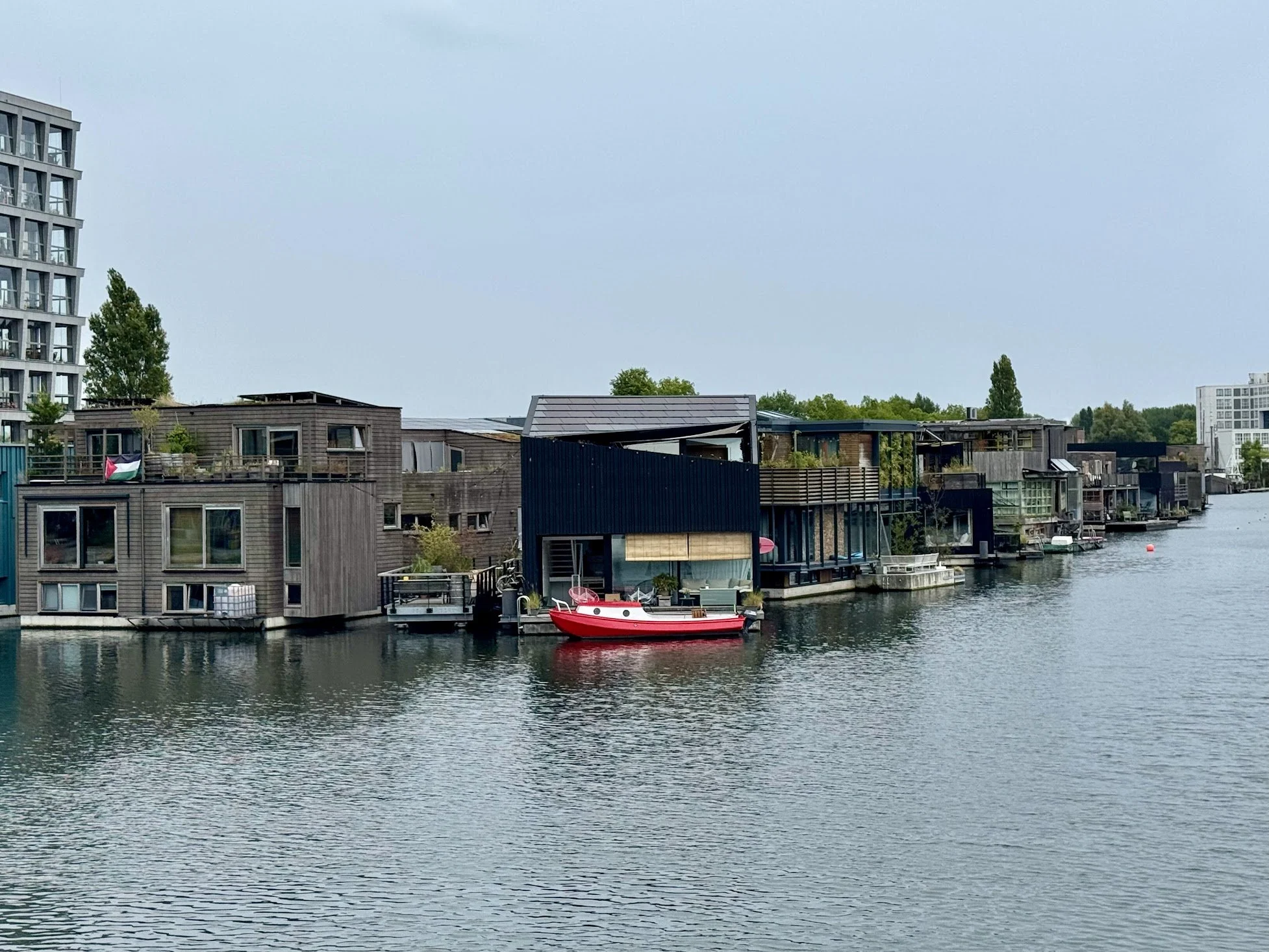 Residential waterfront homes with a red and white boat docked in the water, modern buildings, and trees under a cloudy sky.