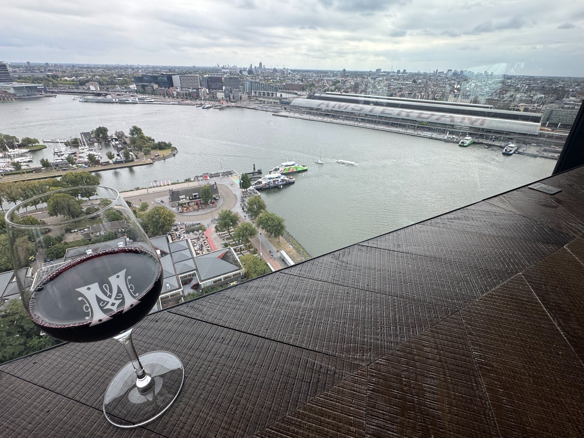 A nighttime city view from a high-rise window, showing tall buildings, water with boats, and a cloudy sky. In the foreground, there is a glass of red wine on a wooden table, partially visible.