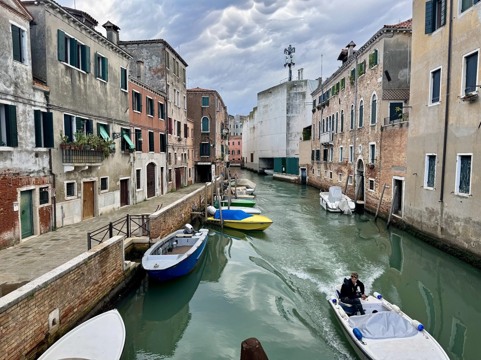 A canal in Venice, Italy, lined by old buildings with colorful facades, some with plants on balconies. Several small boats are docked along the canal, with one boat carrying a person. The sky is cloudy, and the water reflects the buildings.