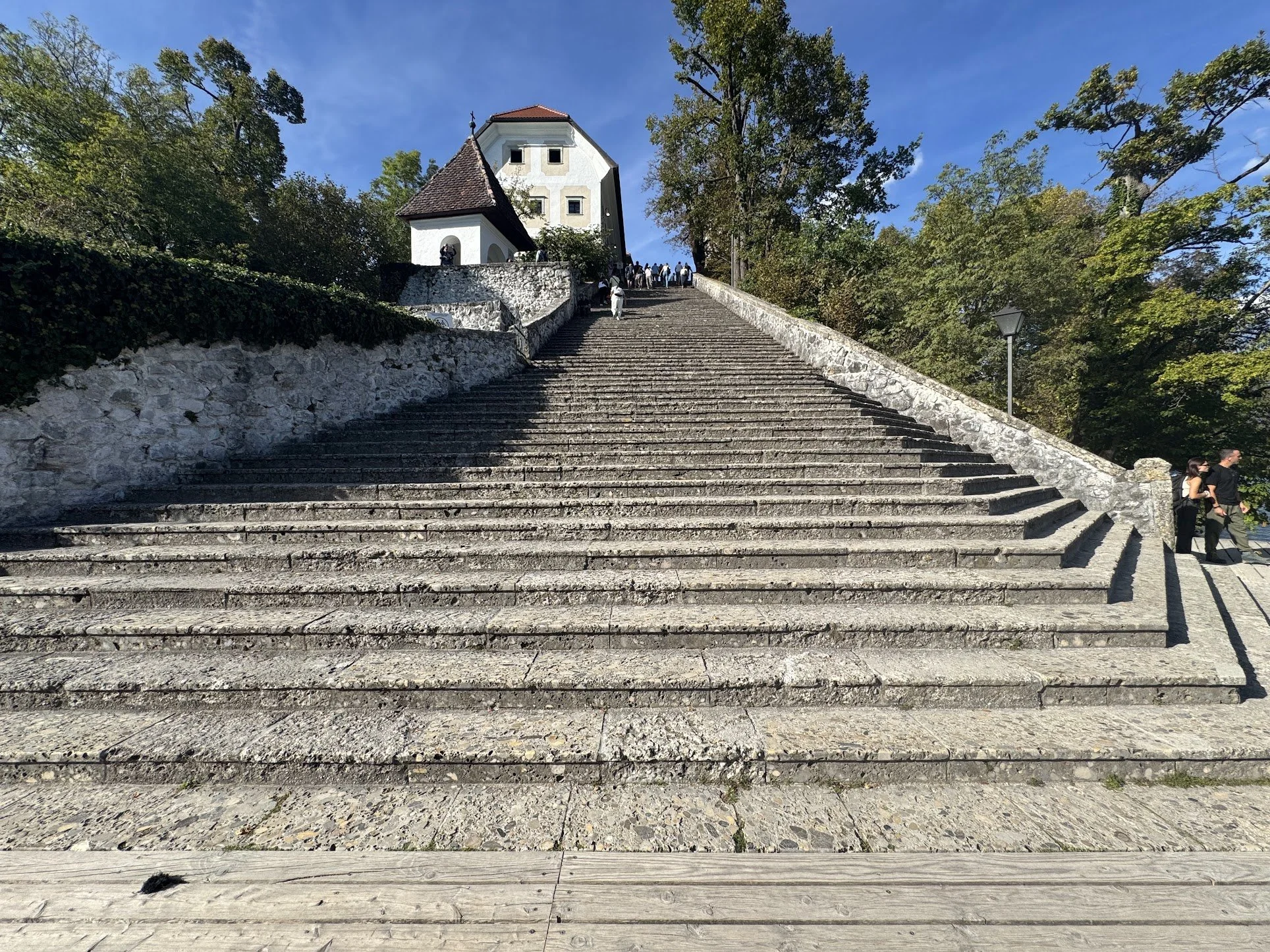 Steep stone staircase leading up to a small white chapel at the top, surrounded by green trees, with people walking up and down the stairs on a sunny day.