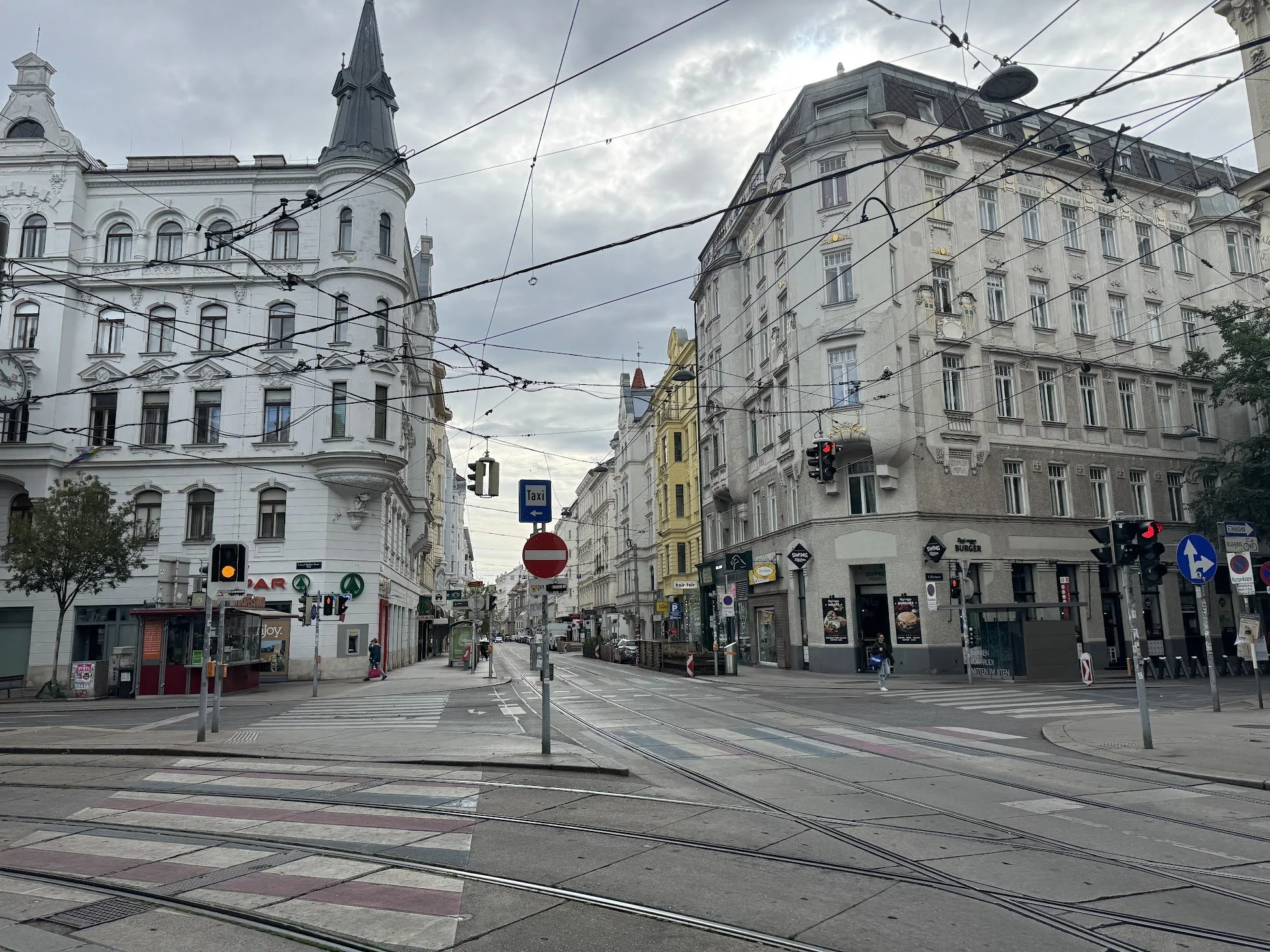 A city street with European-style buildings, tram tracks, traffic lights, and overhead trolley wires under a cloudy sky.
