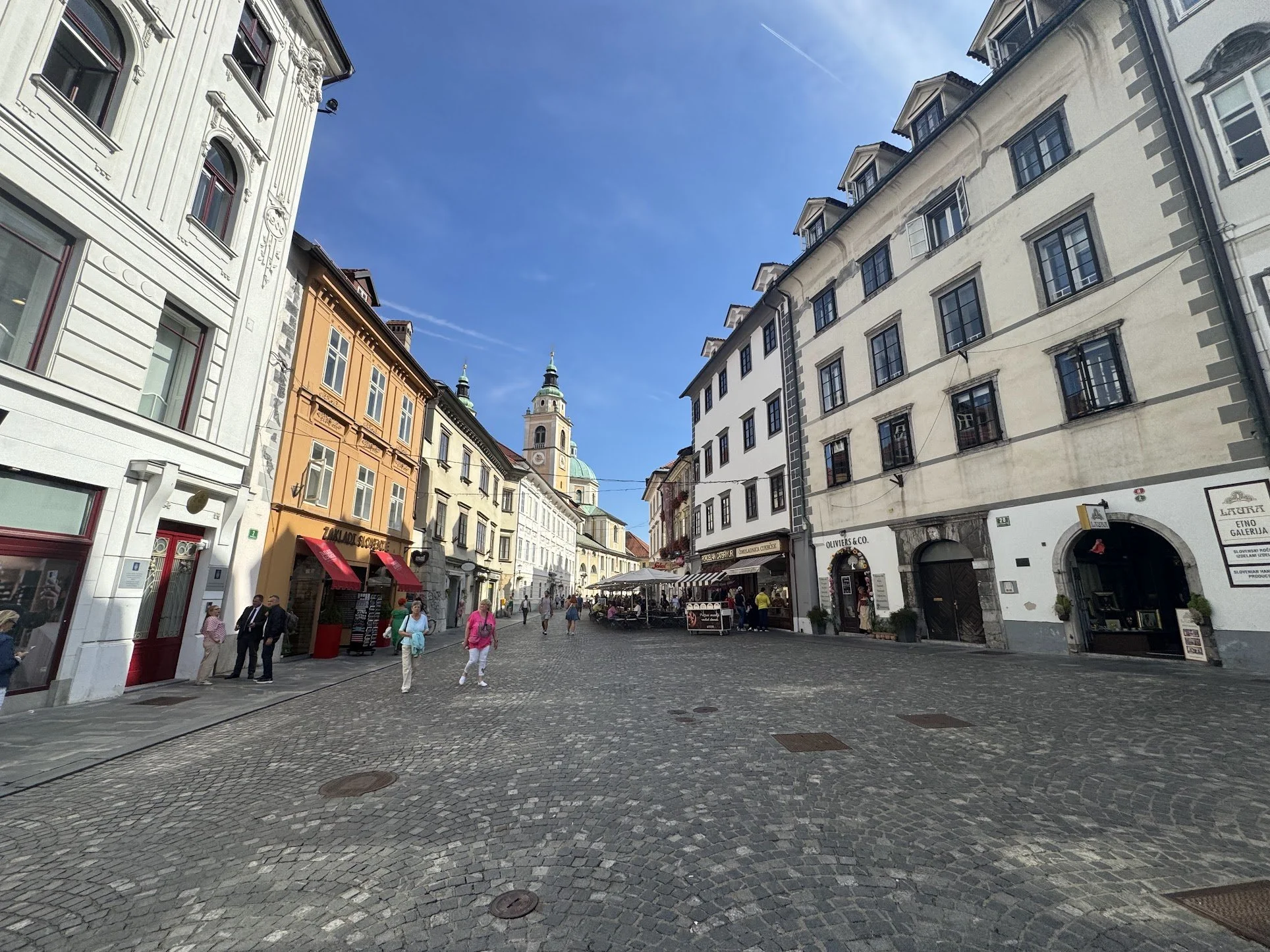 Street view of a European city with historic buildings, people walking on cobblestone street, and a church with a green dome in the background.