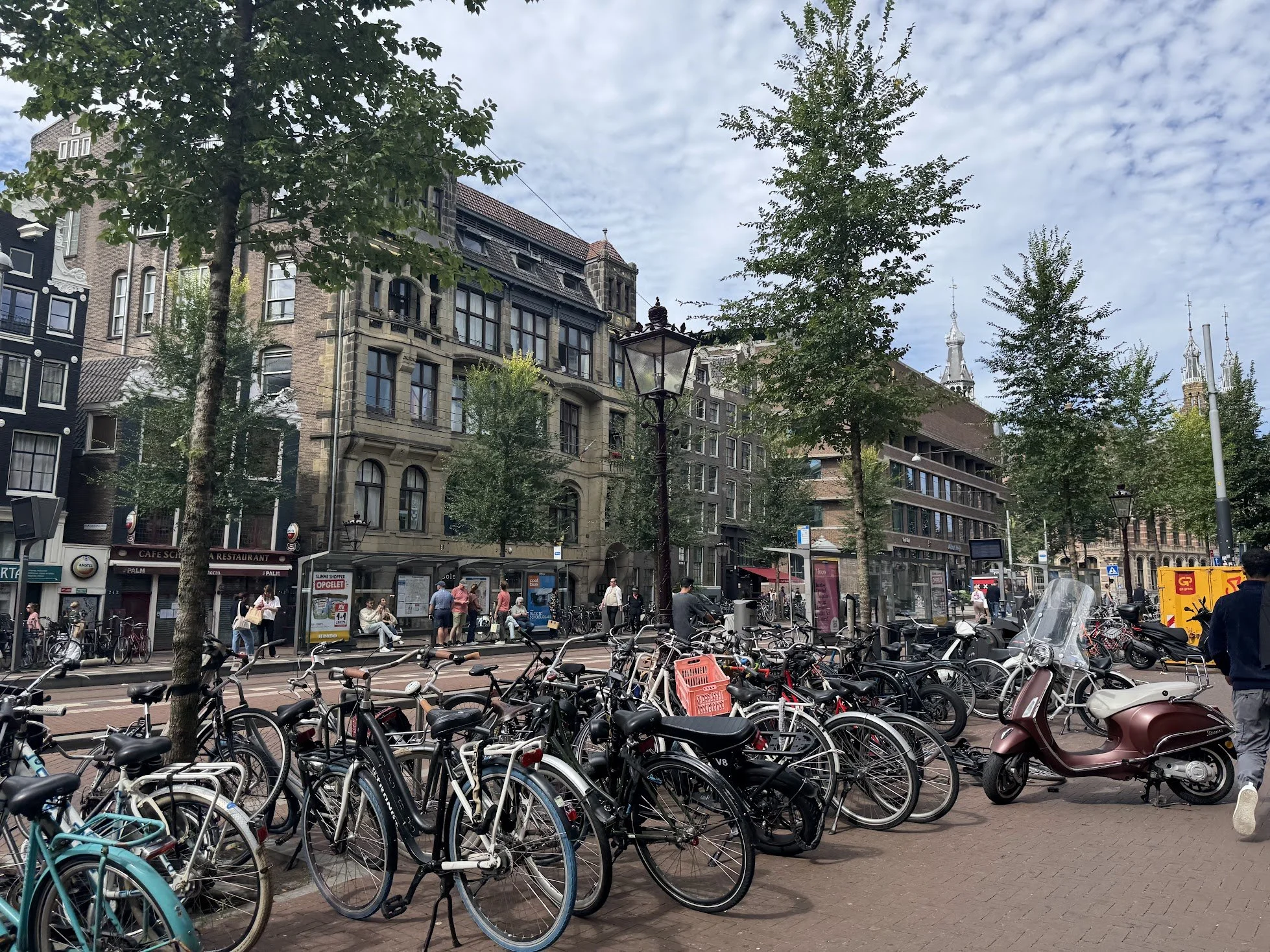 Bicycles and scooters parked on the street in front of a busy city square with trees, historic buildings, and a few people walking or sitting.