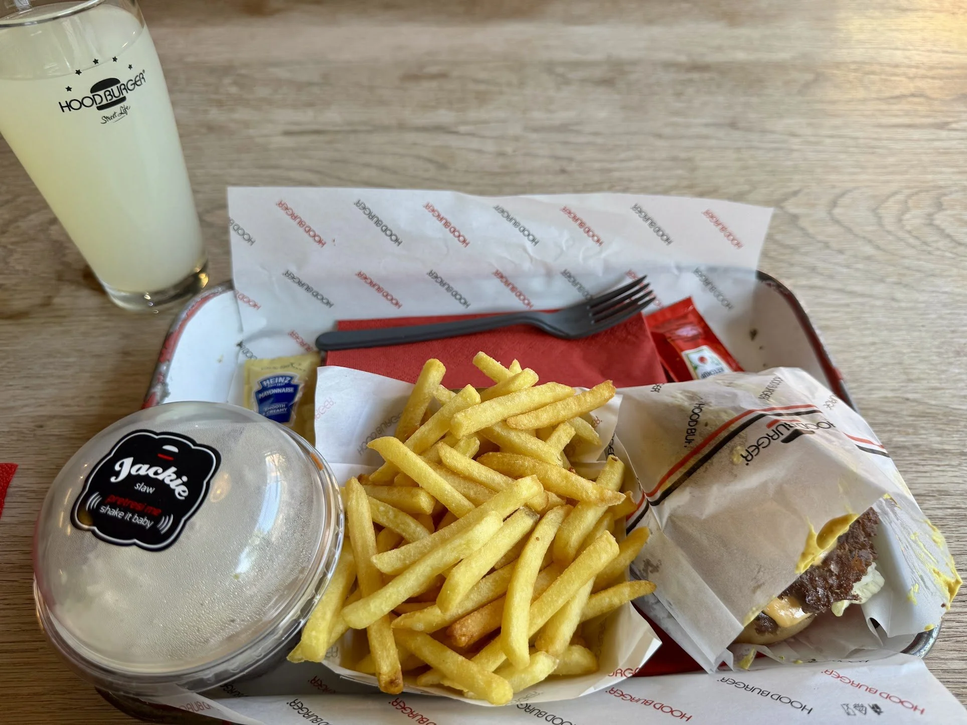 Tray with French fries, a sandwich wrapped in paper, a small container of mayonnaise, a burger in a bun with cheese and bacon, a fork, a paper napkin, and a glass of lemonade at a fast-food restaurant.